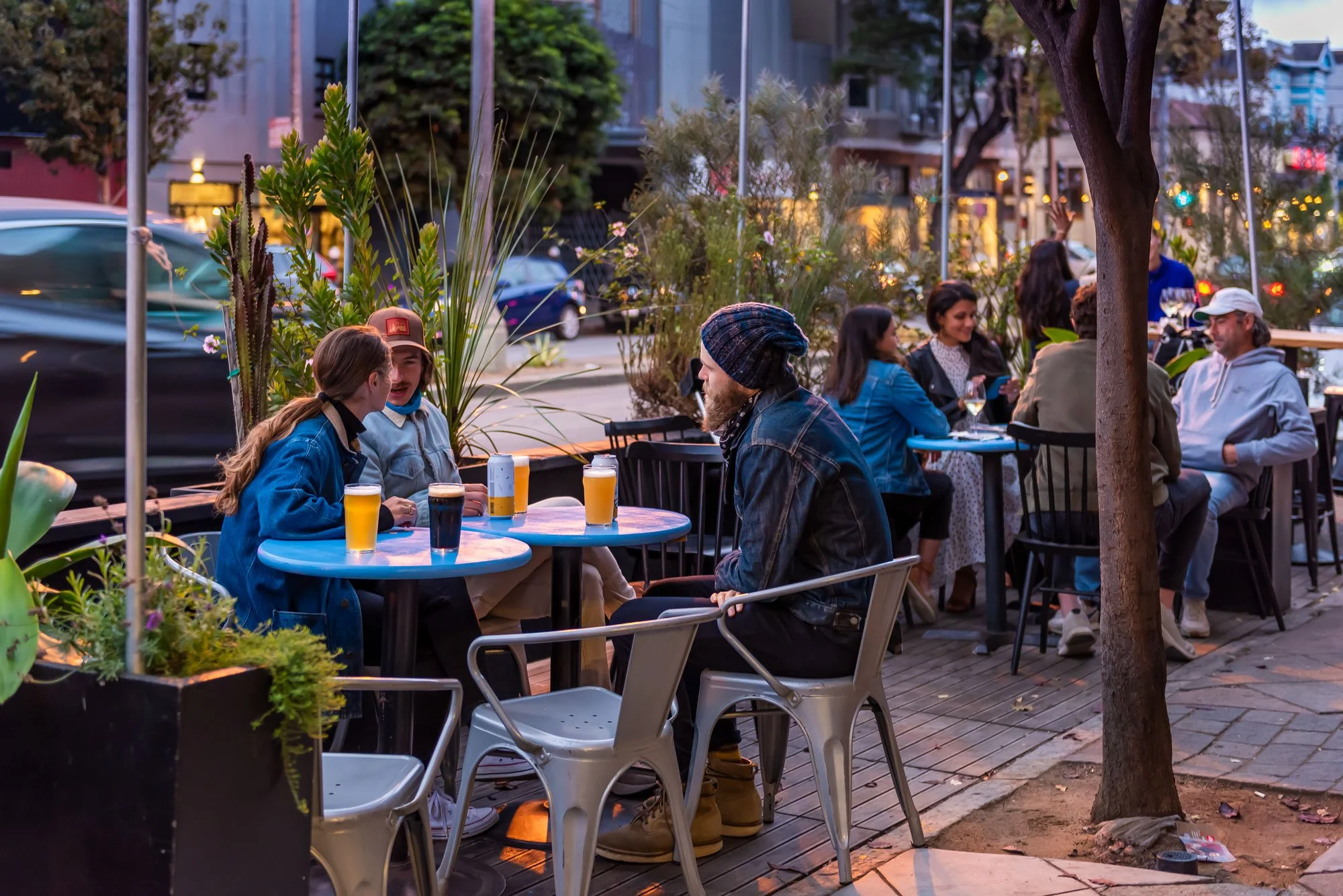 People dining at outdoor restaurant on a city street in the evening, with drinks and conversations among groups of friends.