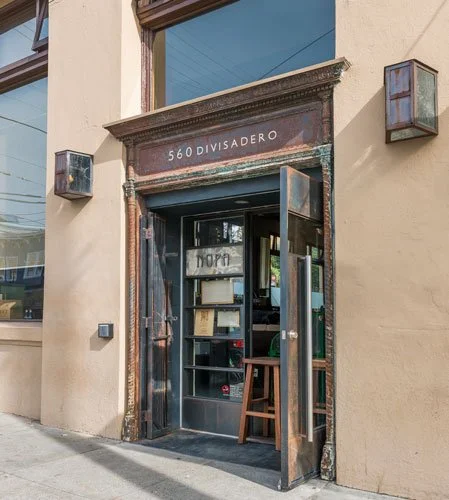 The entrance to a building with a rusted metal door frame, open doors, and the address 560 Divisadero displayed above. Inside, books and a small wooden stool are visible.