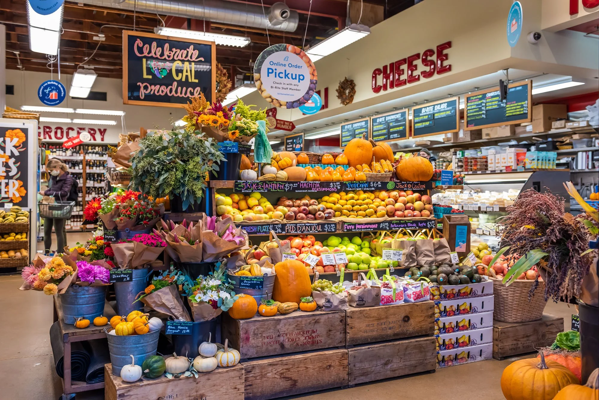 Fresh produce and flowers arranged on wooden crates and metal buckets inside a grocery store, with pumpkins and apples displayed prominently, and signs indicating produce names and prices.