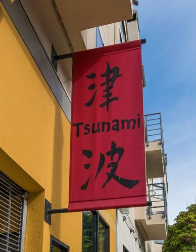 A red vertical banner with black Japanese characters and the word 'Tsunami' written in English, hanging outside a building with a yellow wall.