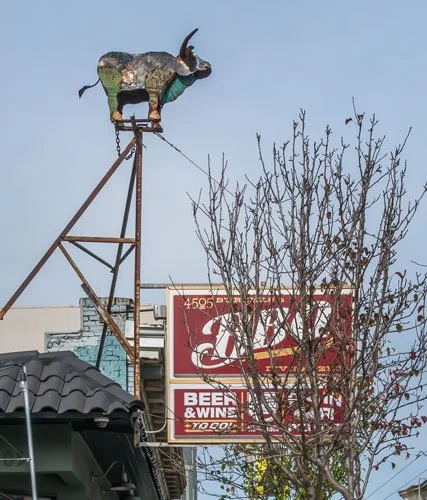 Rusty goat sculpture on a tall metal structure above a building and a sign for a bar and grill.