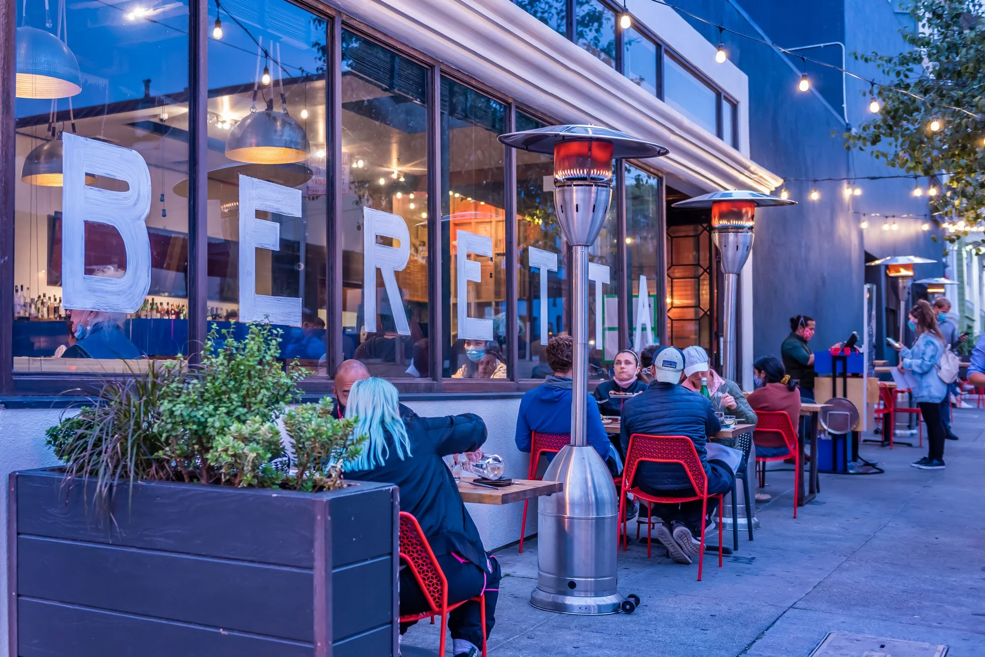 People dining outside at a restaurant with large windows, string lights, patio heaters, and plants, during evening.