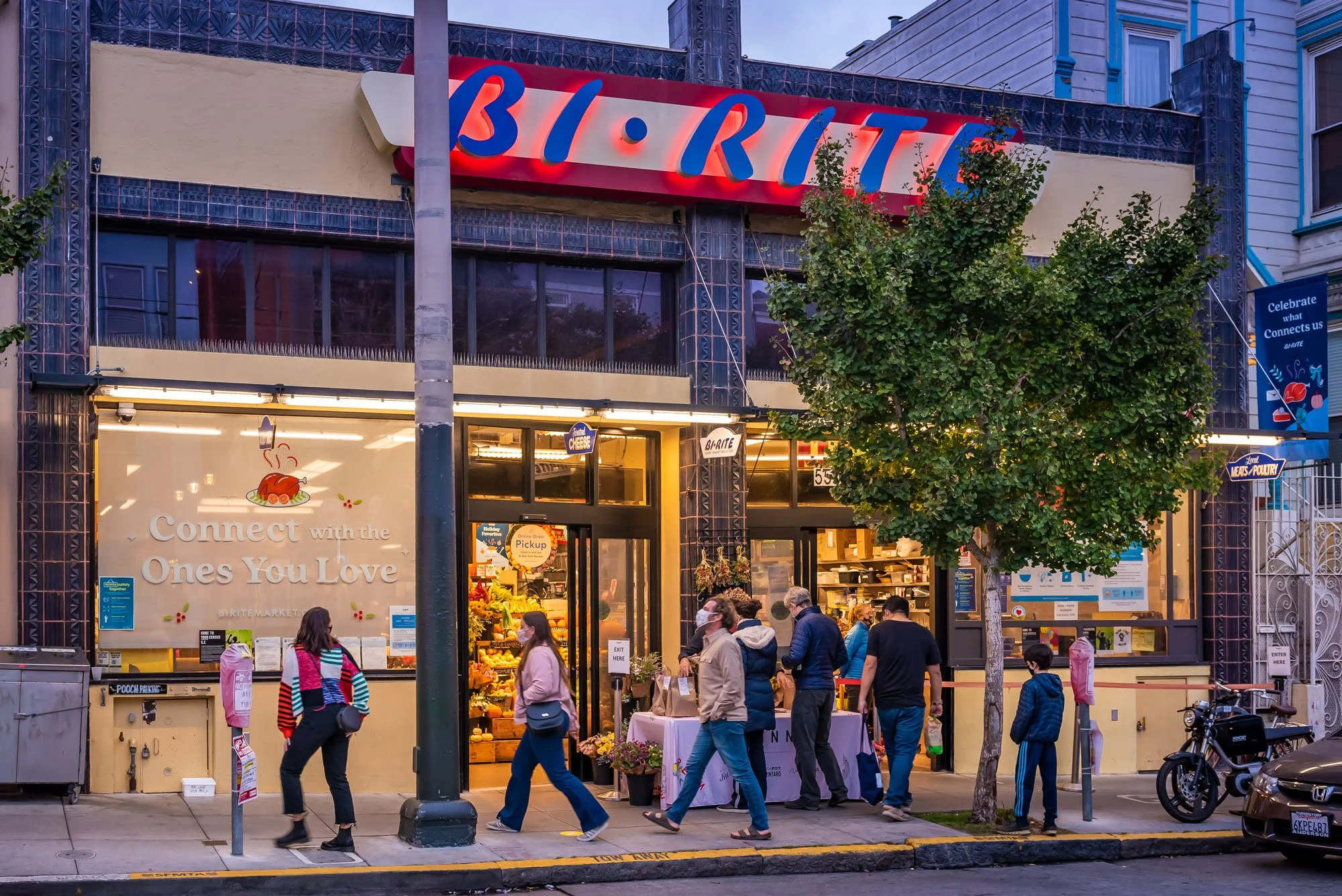People entering and leaving a busy grocery store with a large sign that reads 'BIRITE' above the entrance, a tree in front, and a sidewalk with a few parked bikes and cars.