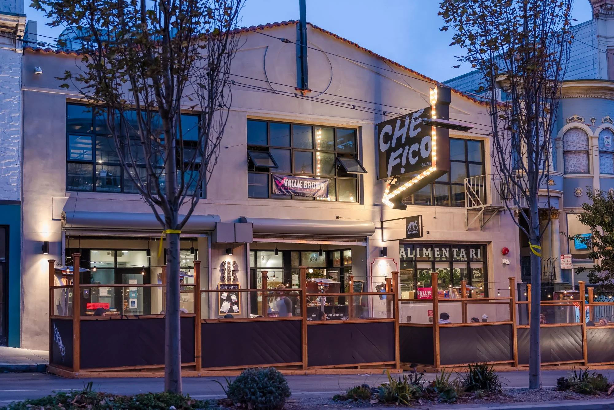 Exterior of a restaurant named Che Fico with outdoor seating, illuminated sign, and large windows, during dusk in an urban setting.
