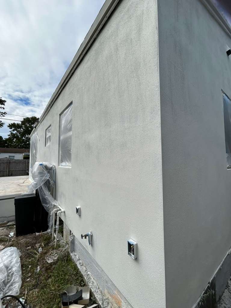 A black wooden pergola under construction with a ladder and tools around, surrounded by tropical palm trees and a fence, with a partly cloudy blue sky.