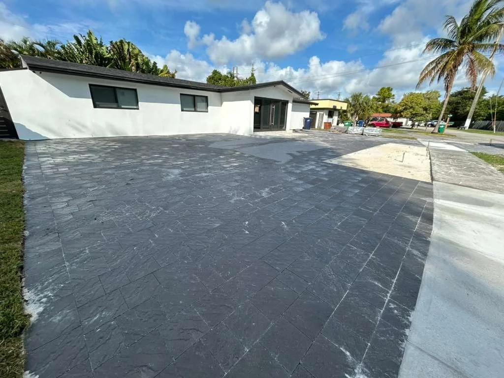 Construction site with workers installing gray brick pavers on a sidewalk, surrounded by tropical trees and buildings in the background.