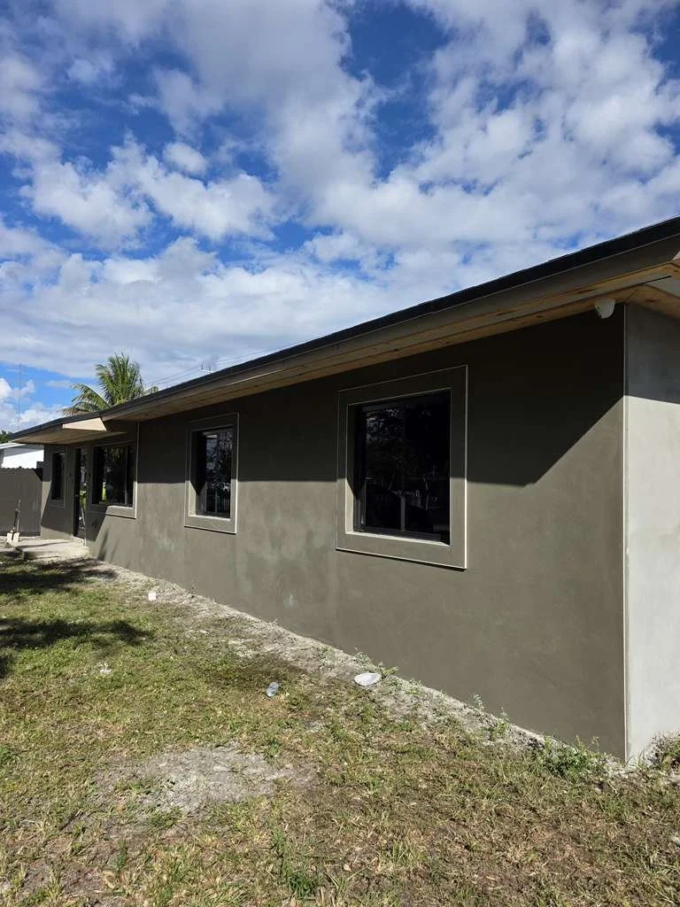 Side of a building with a gray exterior wall and white gutter, with shadows of tree branches cast on the wall. There is a large tree next to the building, and a cluttered area with construction materials and a pot on a table near the building.