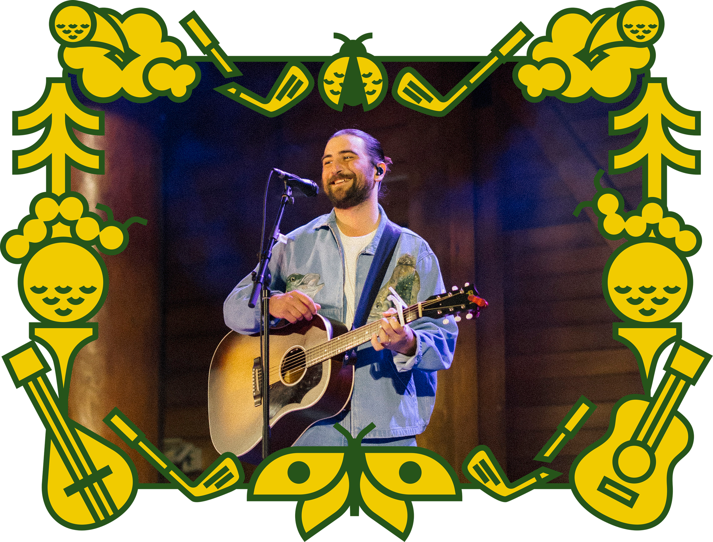 A man with a beard and long hair tied back, smiling and playing an acoustic guitar on stage, with a microphone in front of him.