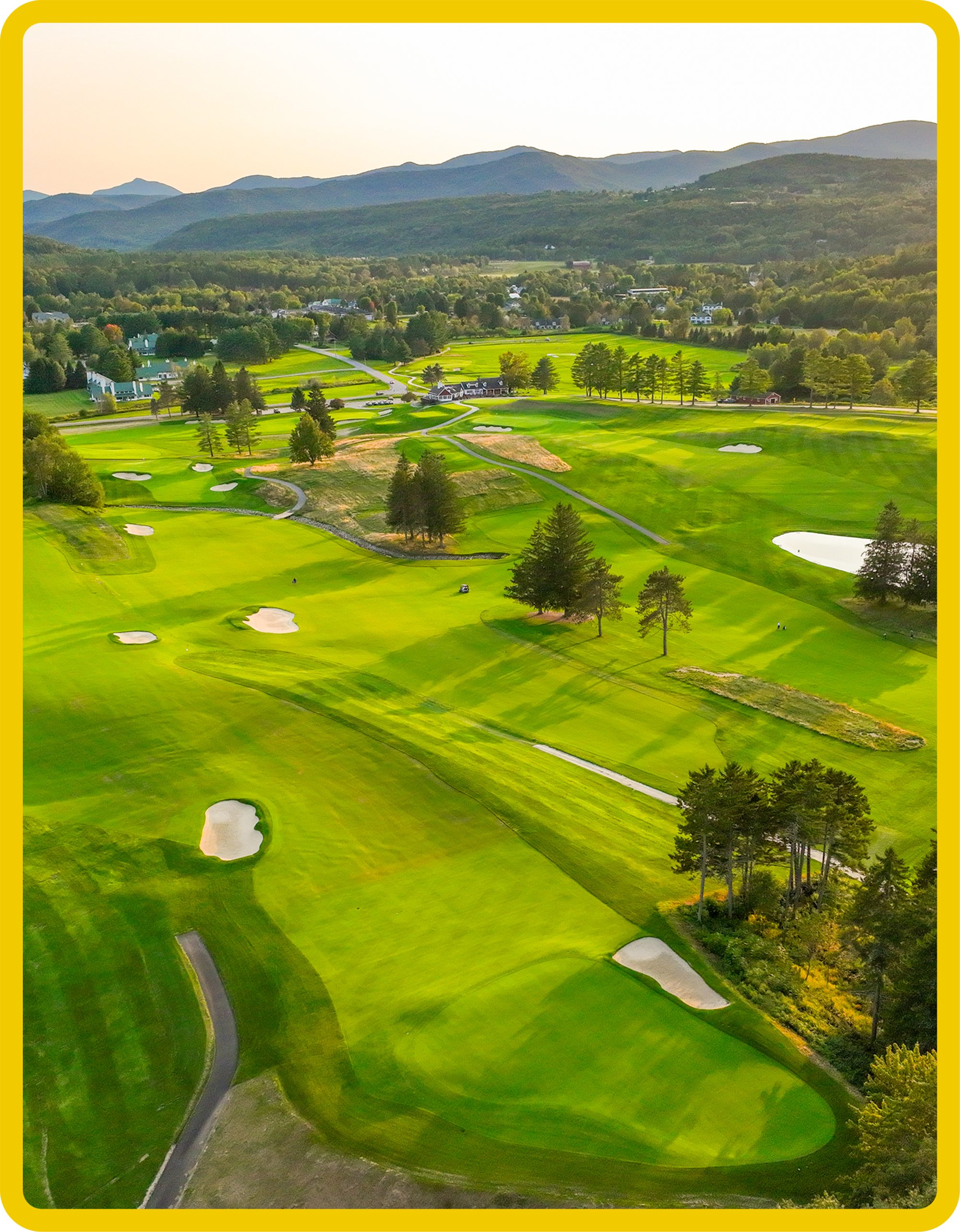Aerial view of a golf course with green fairways, sand traps, scattered trees, and a small building, set against a backdrop of mountains and a clear sky.