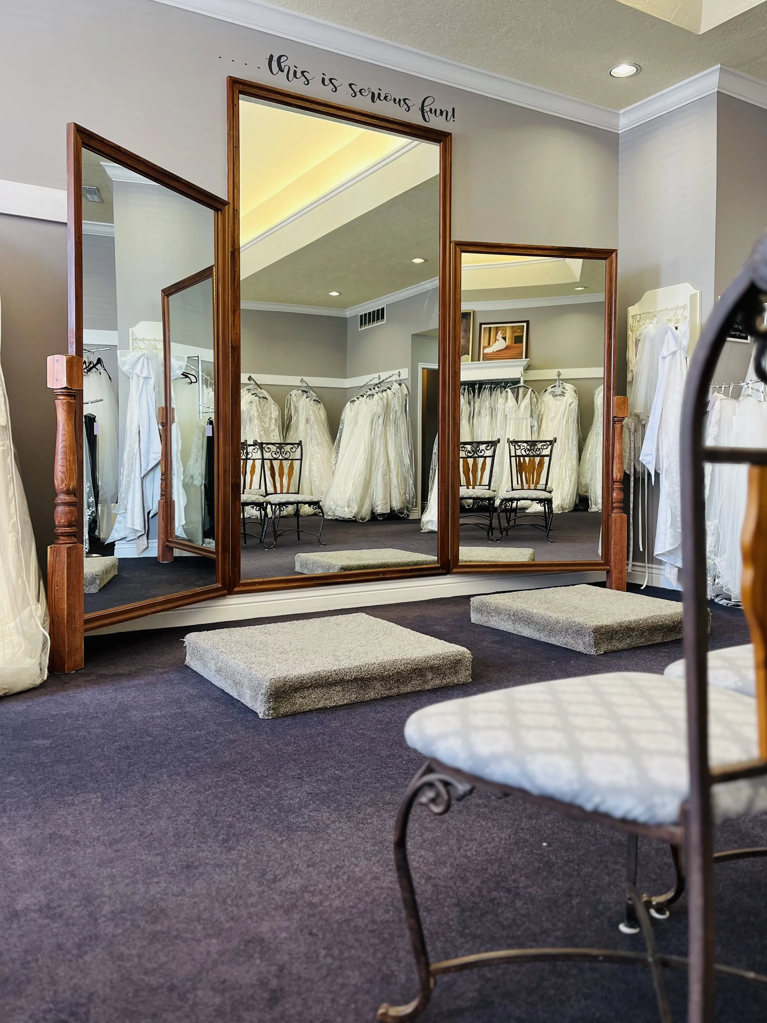 A fitting room with a large mirror, surrounded by wedding dresses on racks in a bridal boutique. There are small carpet steps in front of the mirror for trying on shoes. The wall above the mirror has the phrase 'this is serious fun!' written in black cursive.