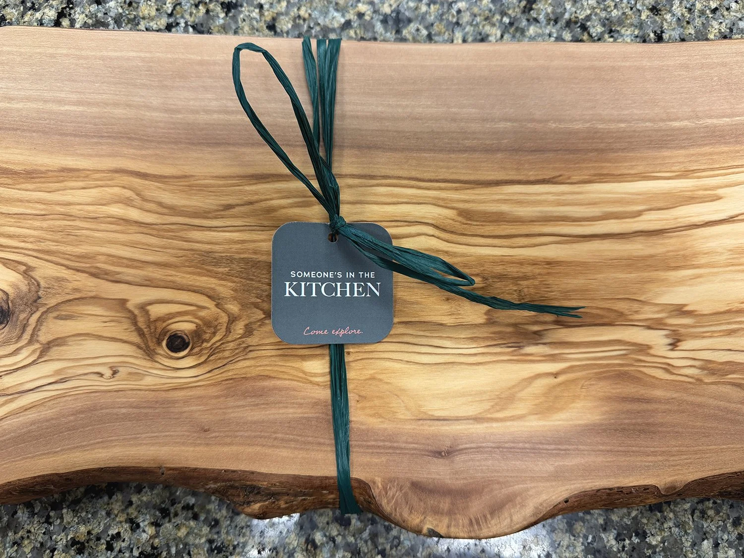 A rectangular piece of wood with a small gray tag attached by a green string. The tag reads 'Someone's in the Kitchen' with the phrase 'Come explore.' underneath.