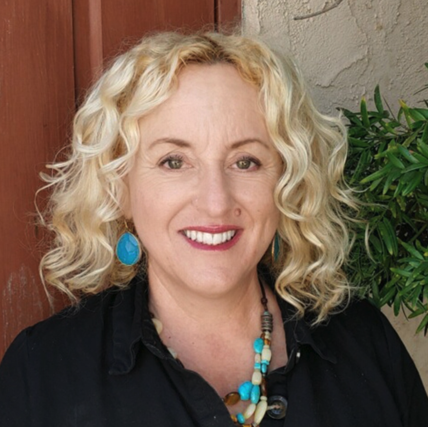 A woman with curly blonde hair, wearing turquoise earrings and layered necklaces, smiling in front of a wooden and stucco wall, with a green plant in the background.