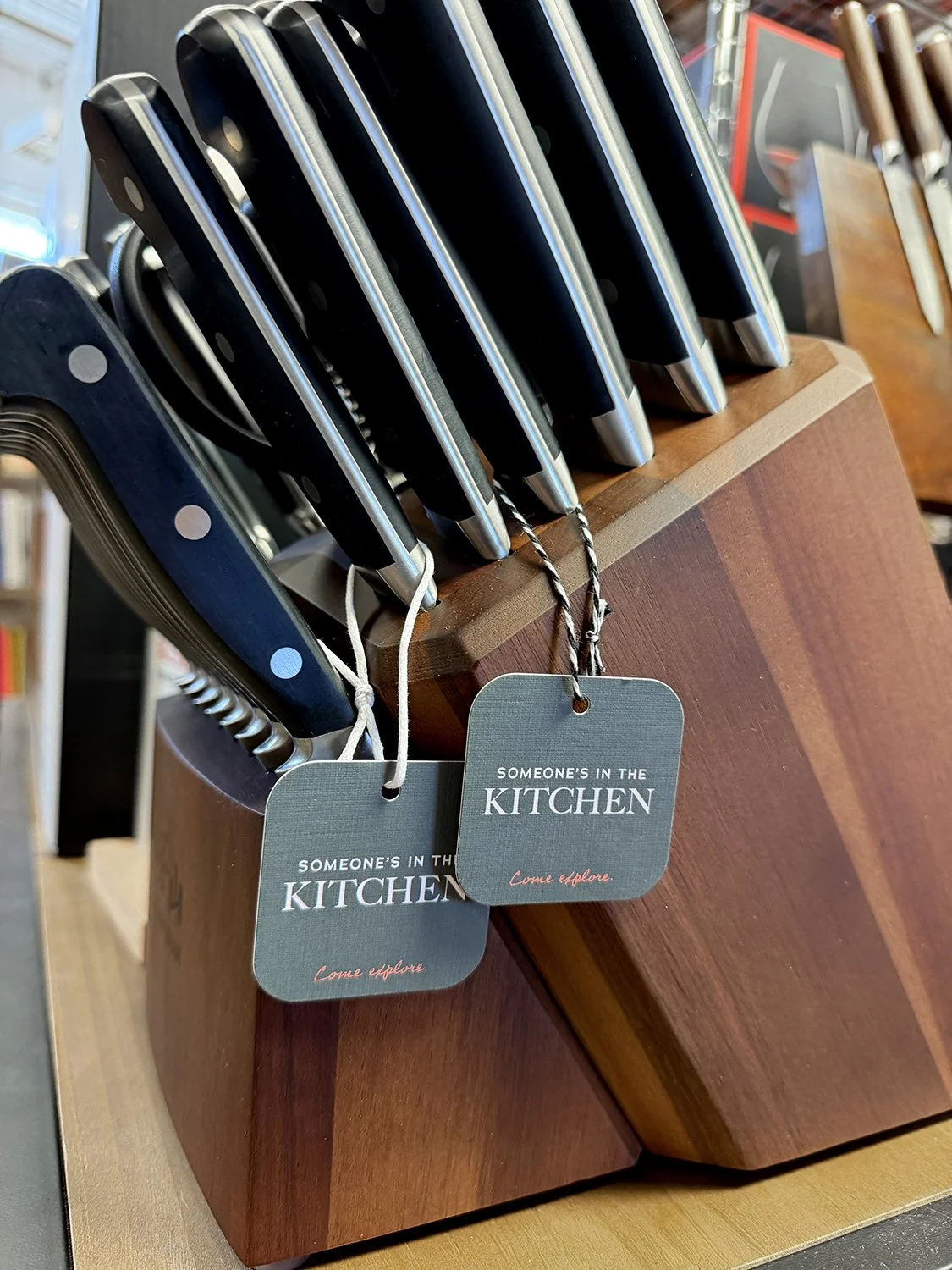 A set of kitchen knives with black handles and stainless steel blades in a wooden knife block, with tags that read 'Someone's in the Kitchen' and 'Come explore'.