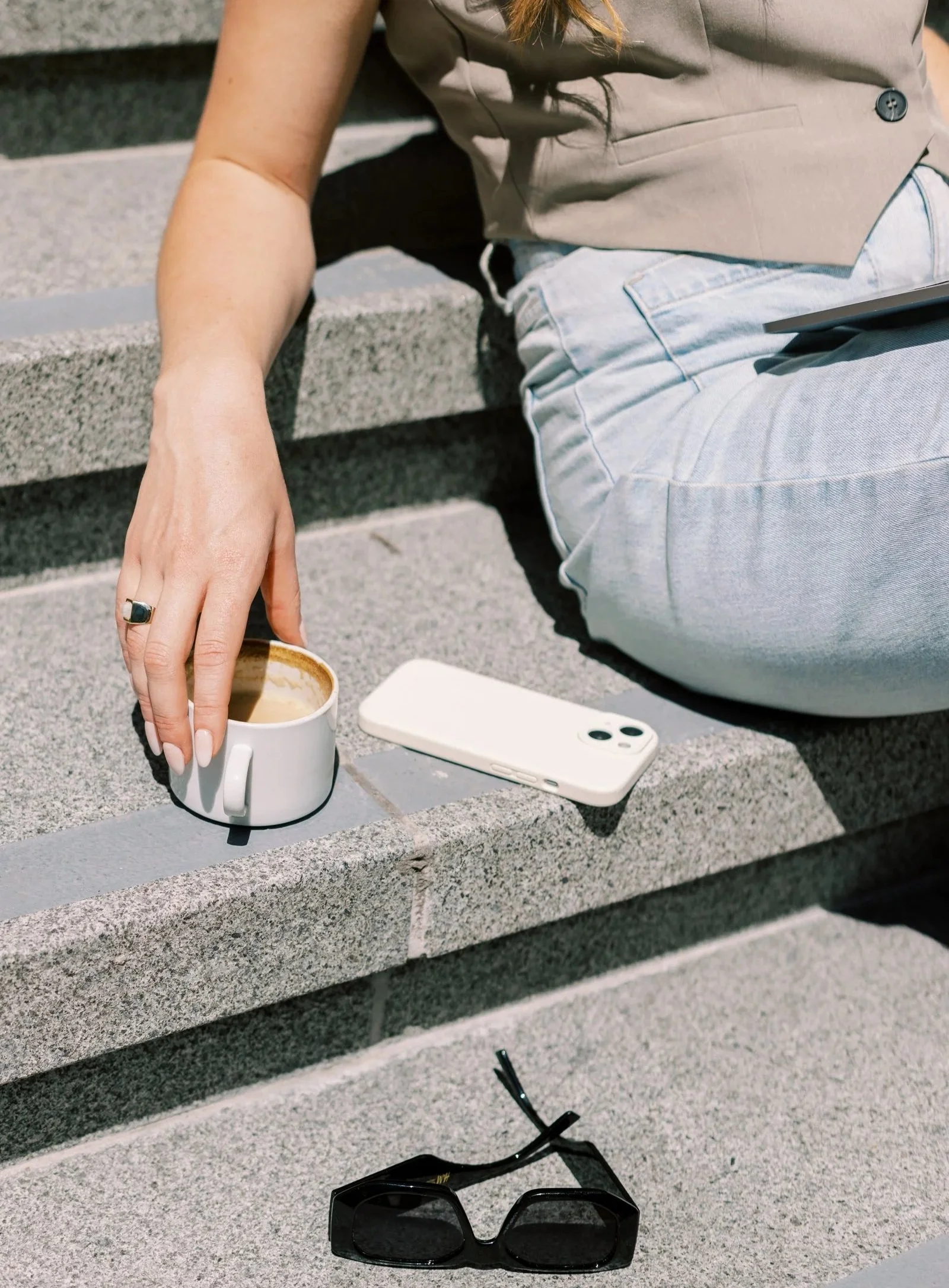 A person sitting on outdoor stairs with a coffee mug, smartphone, and sunglasses placed on the steps near them.