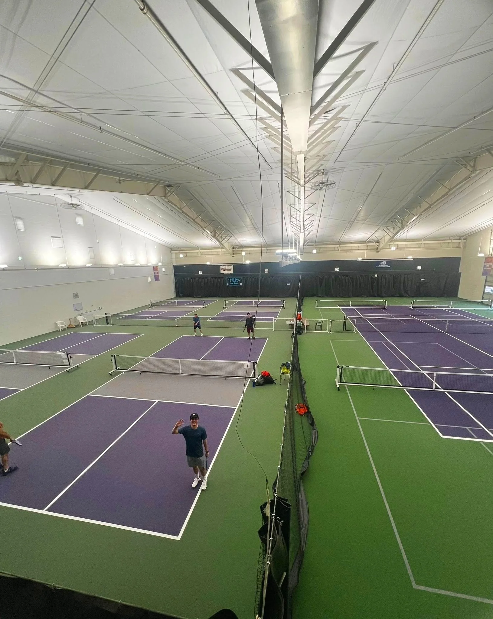 Indoor tennis facility with multiple purple and green courts. Several people are on the courts, and one person in the foreground is waving and smiling at the camera.