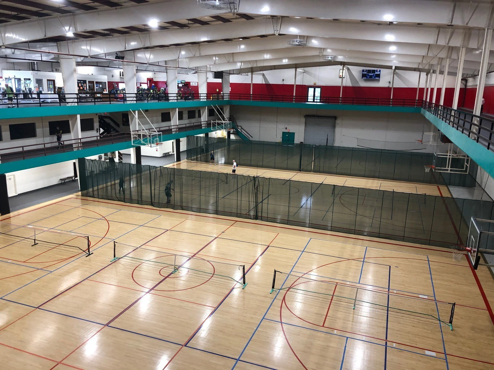 Large indoor sports facility with multiple courts separated by green nets and a basketball court with hoops. The facility has a high, arched ceiling and a second-floor balcony with a black railing, overlooking the courts.