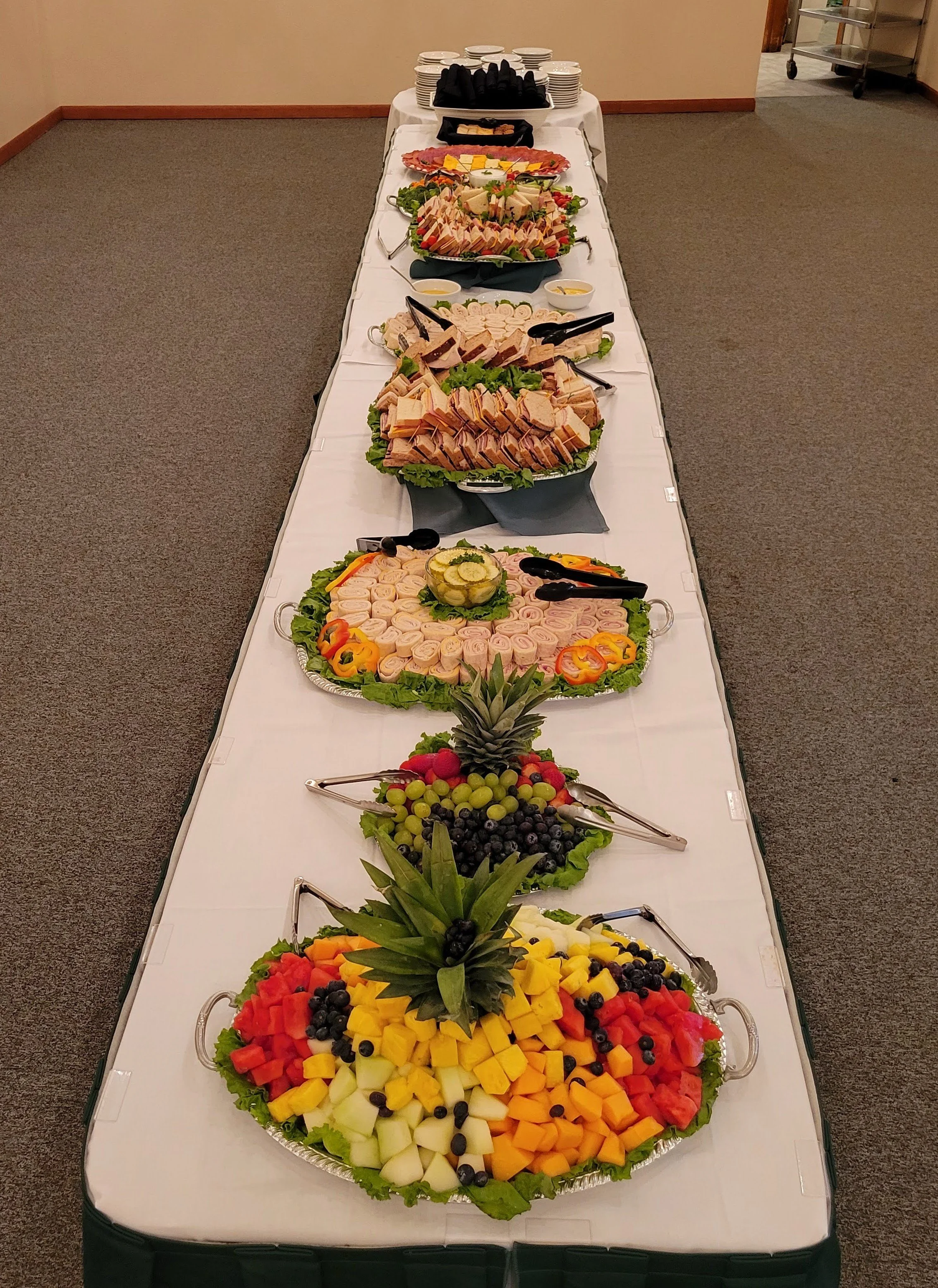 A long table covered with a white tablecloth displaying a variety of fruit and meat platters, including pineapple, grapes, watermelon, cantaloupe, cheese, and cold cuts, with a dessert tray and stacked plates at the end.