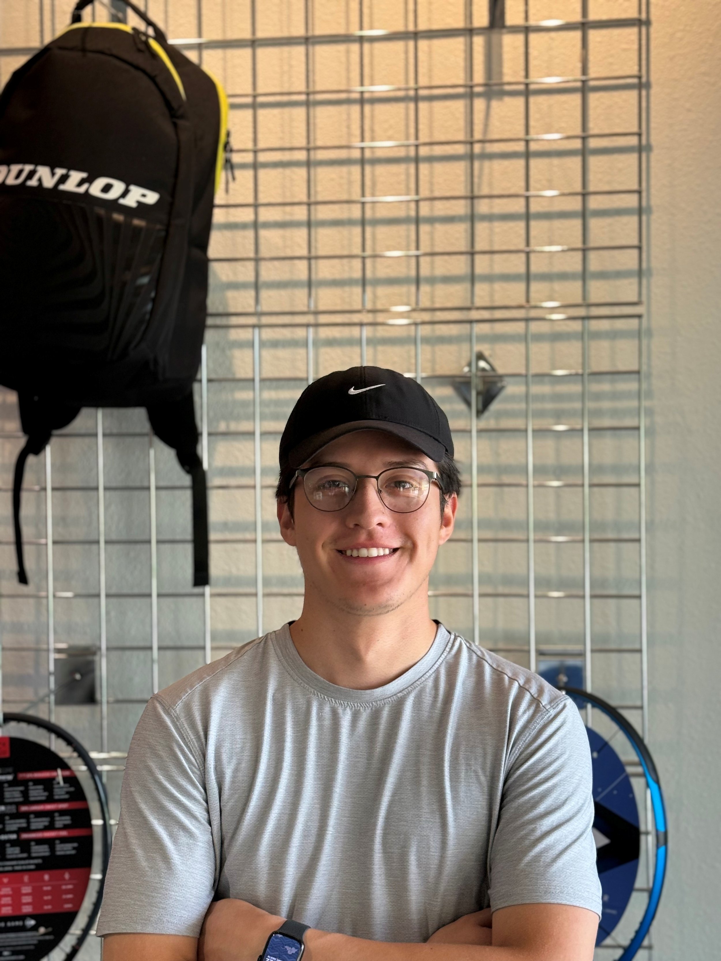 A young man with glasses and a black cap smiling while standing with his arms crossed in front of a metal grid wall with sports equipment, including a black backpack and a blue wheel, hanging on it.