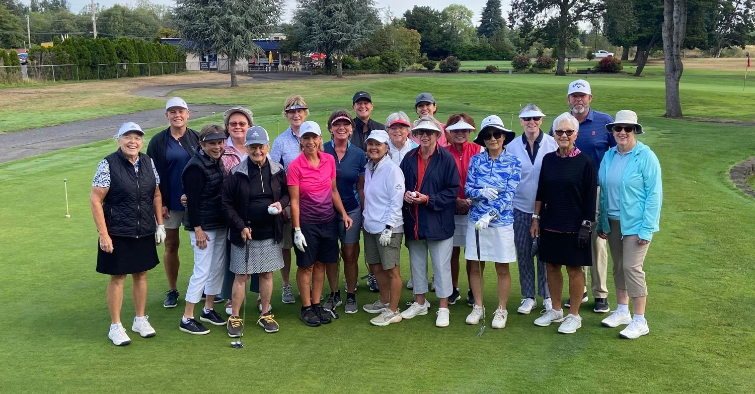 Group of women and men smiling on a golf course, holding golf clubs, wearing casual golf attire and hats, with trees and a clubhouse in the background.