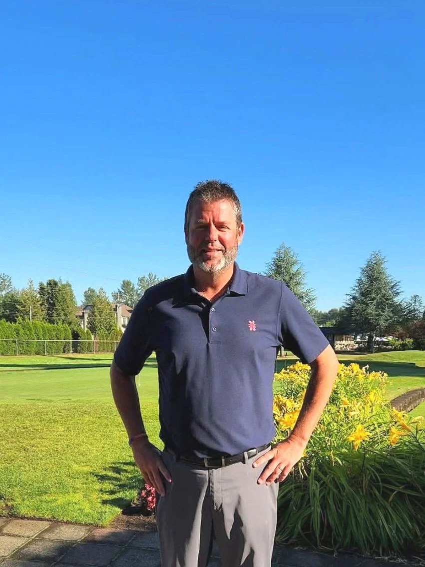 A man standing outdoors on a sunny day, wearing a navy polo shirt and gray pants, with a green park and trees in the background.