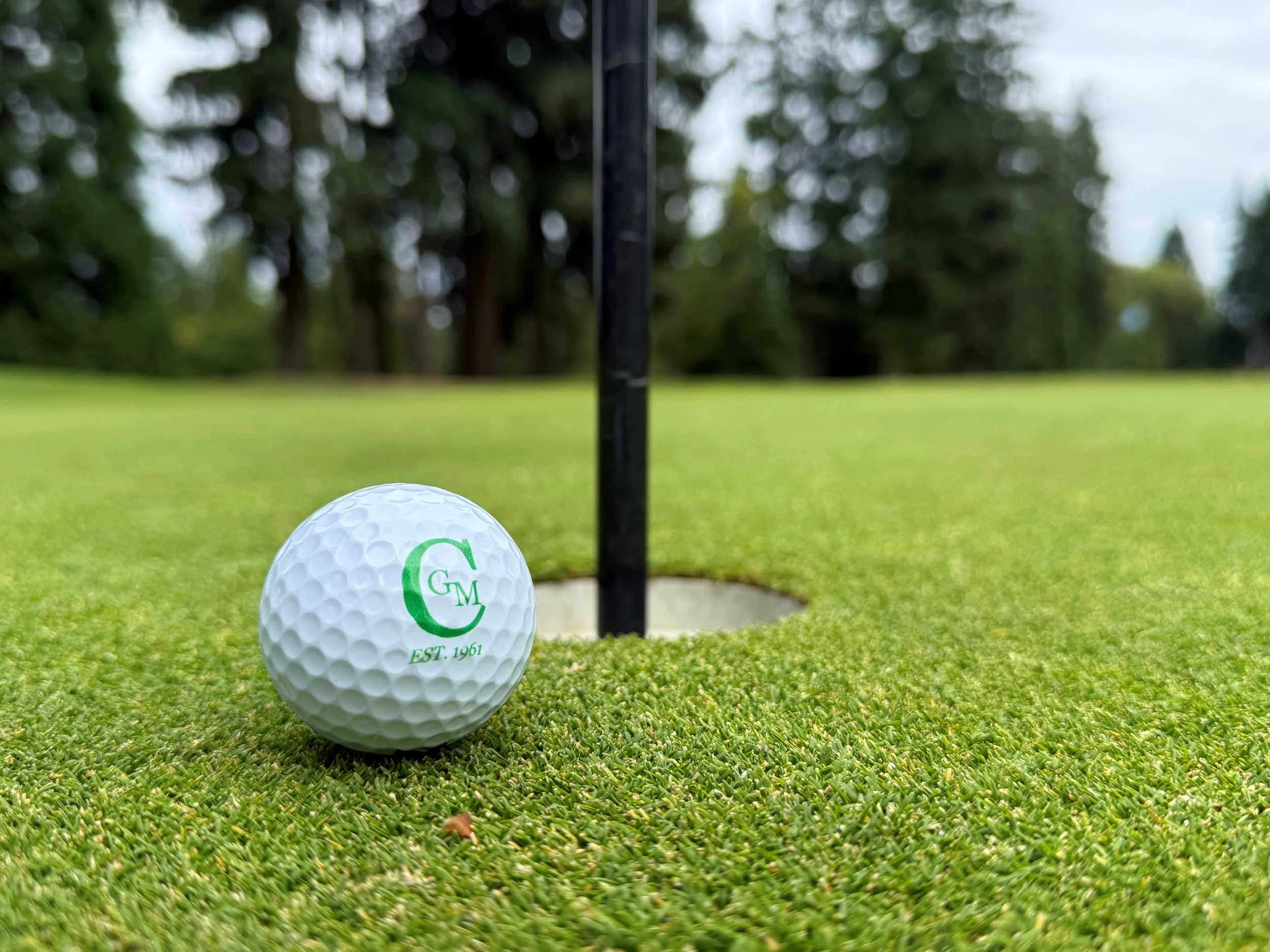 Golf ball with a green logo and the text "EST. 1961" near a hole on a golf green, with trees in the background.
