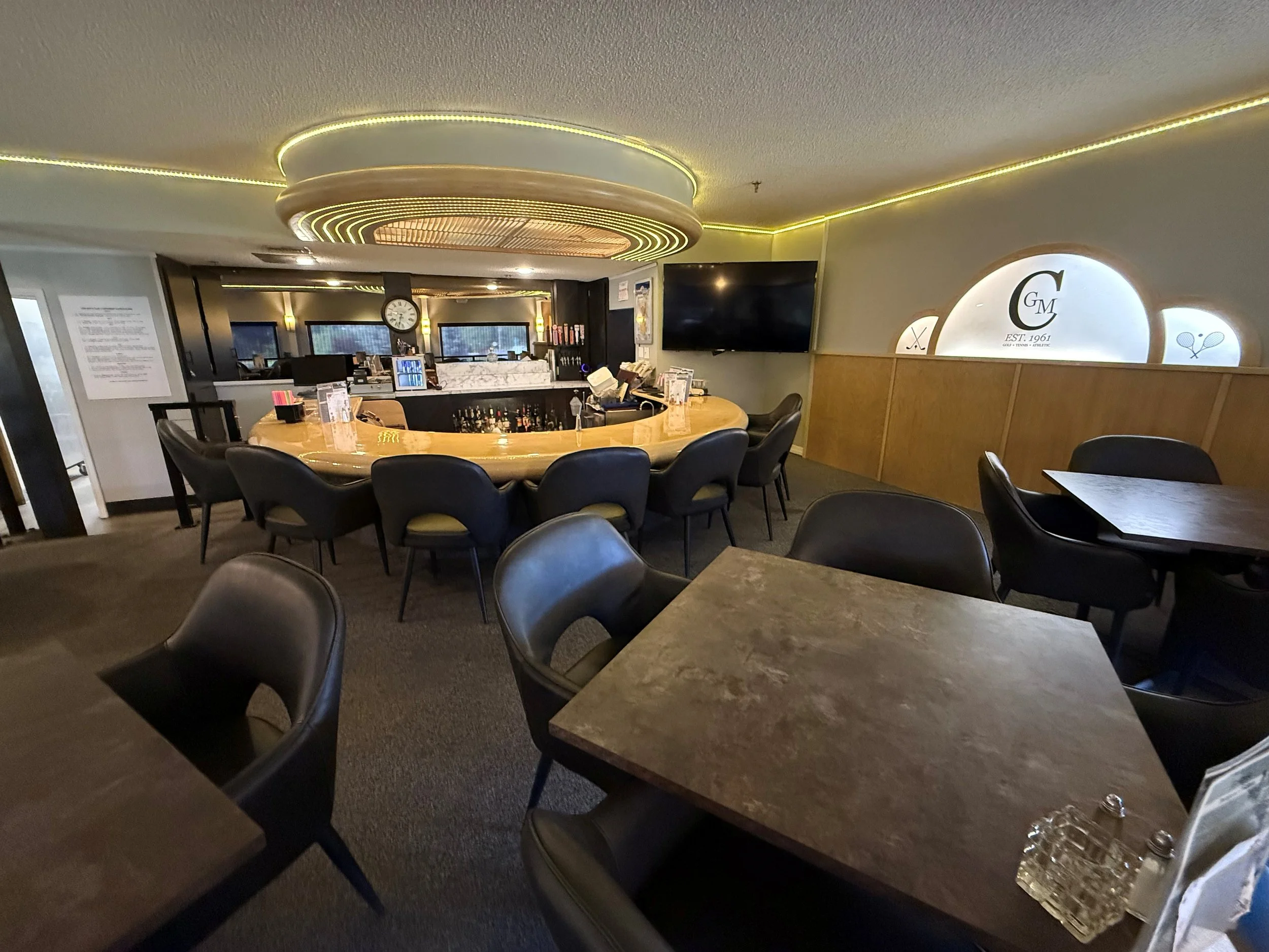 Interior of a bar or restaurant with a curved bar counter, black chairs, a television on the wall, and a decorative illuminated wall sign with the initials 'G M' and