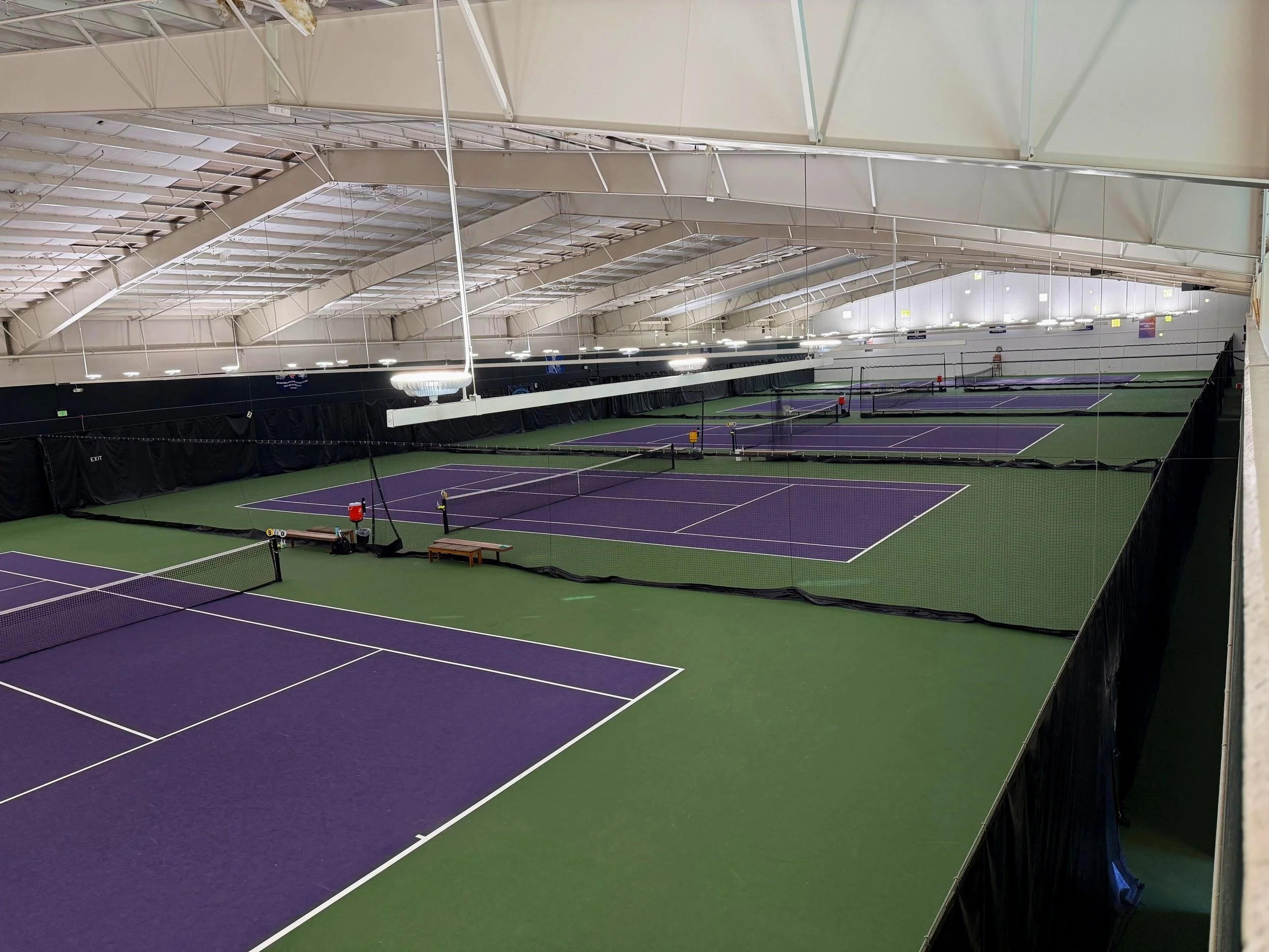 Indoor tennis courts with purple surfaces, nets, and seating areas, under a high arched ceiling with lighting.