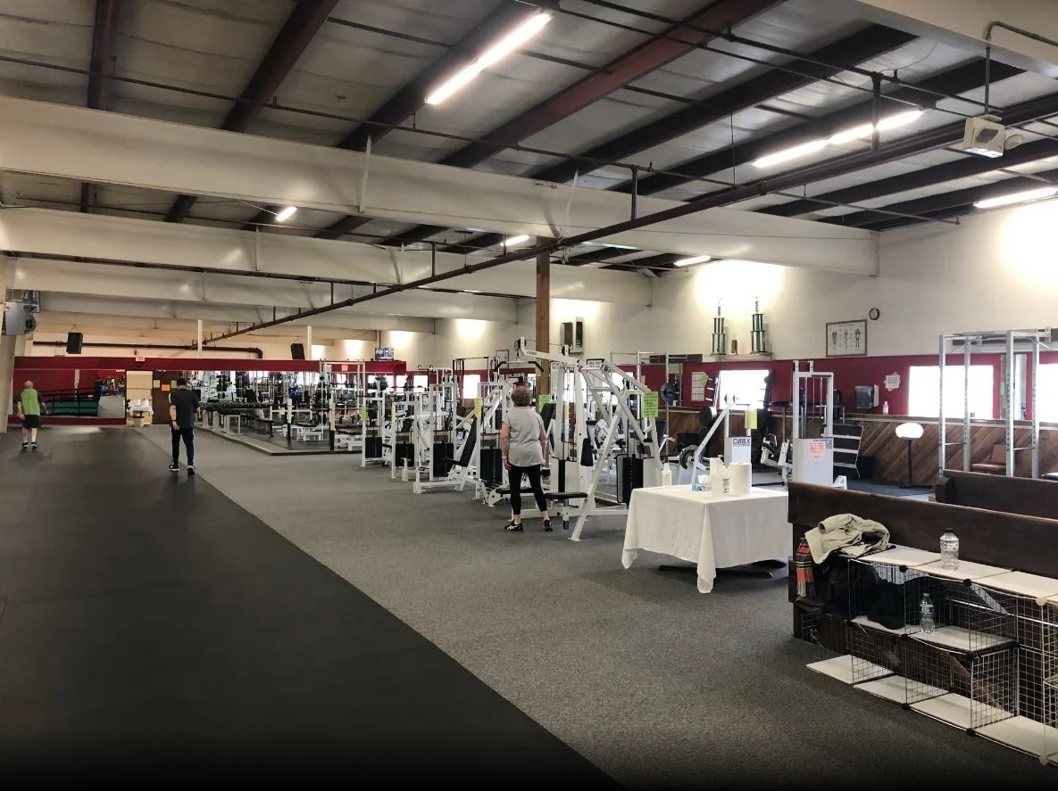 Empty gym with weight machines, people walking, and tables with supplies inside a large, industrial-style fitness center.