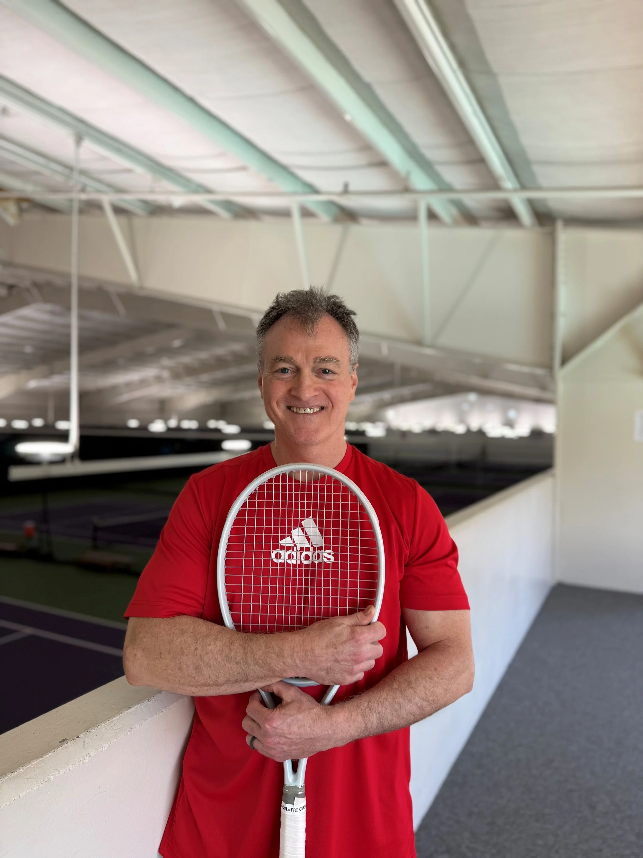 A man in a red shirt holding a tennis racket and smiling, standing on an indoor tennis court.