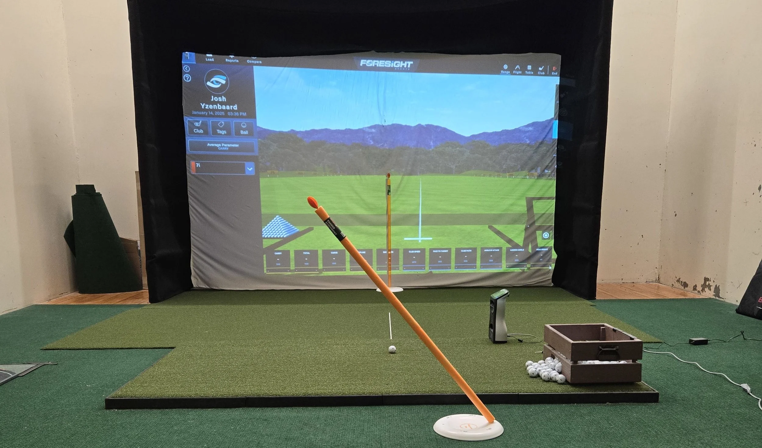 Indoor golf simulation room with a hitting mat, golf ball on a tee, and a projection screen showing a virtual golf driving range.