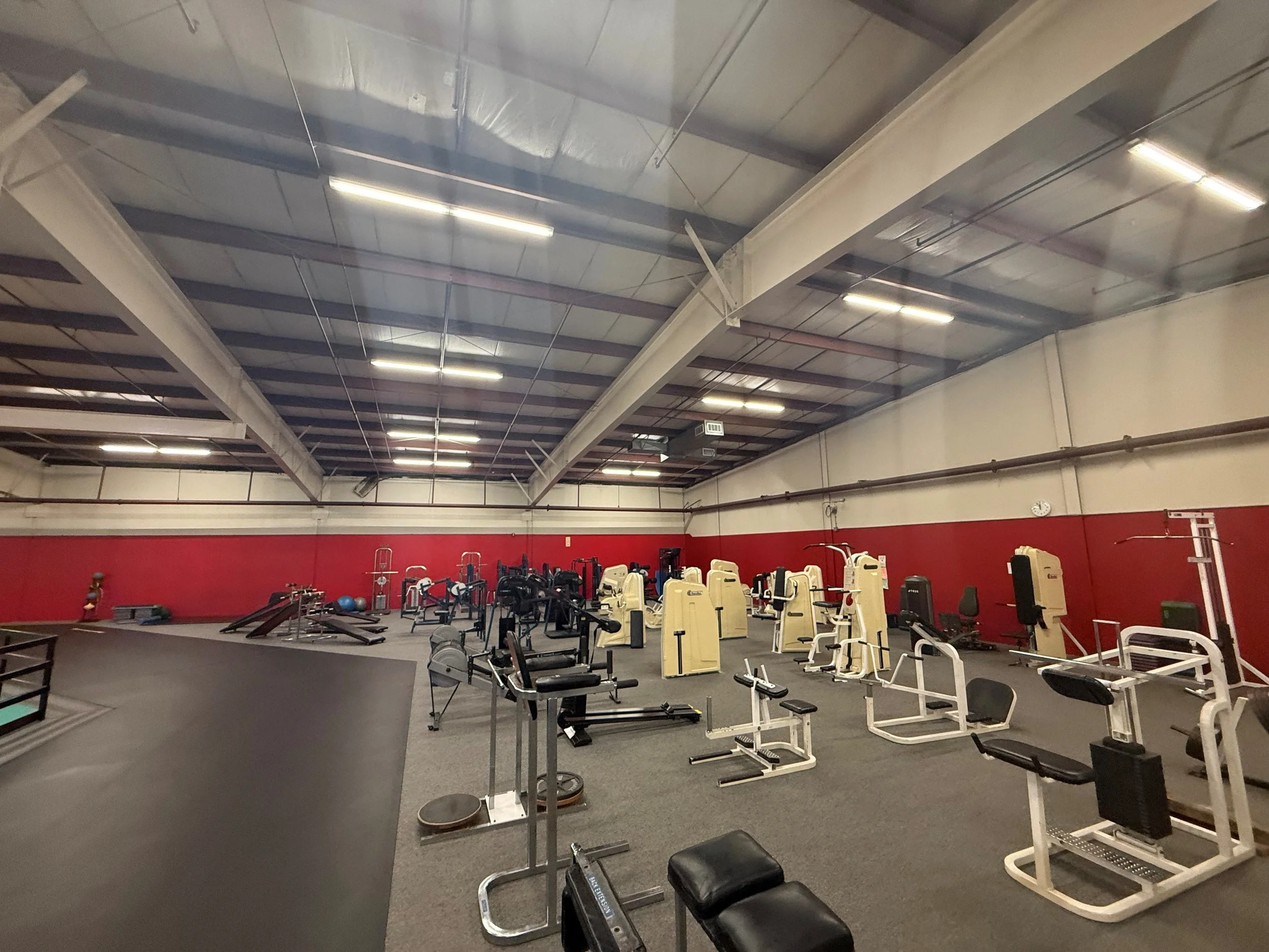 Empty gym with various exercise equipment, including weight machines, benches, and free weights, in a spacious, industrial-style room with a high ceiling and fluorescent lighting.