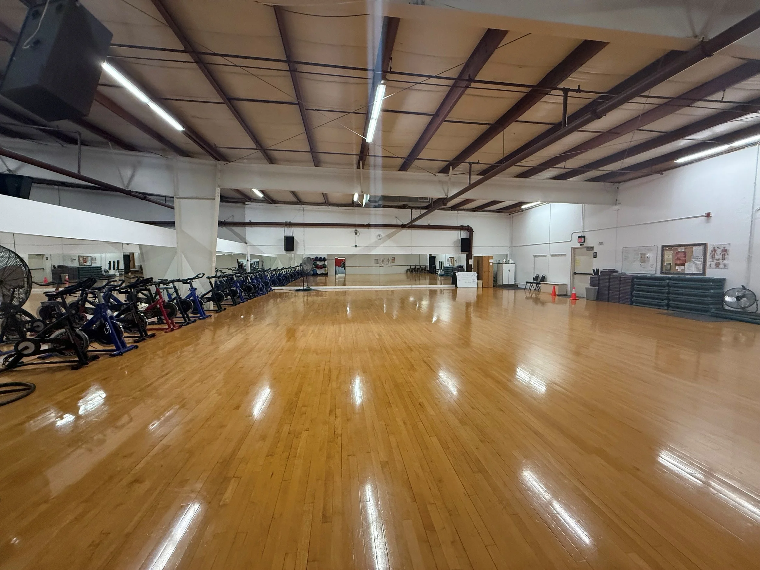 Empty indoor cycling studio with a polished wooden floor, a row of stationary bikes , and fitness equipment against the walls, lights on the ceiling, and mirrors.