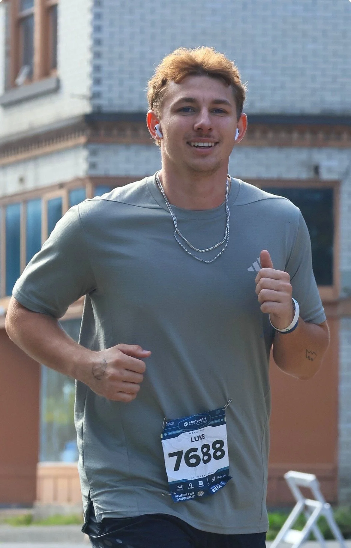 Young man running in a marathon, wearing a gray athletic shirt, white earbuds, a race bib numbered 7688 with the name Luke, necklaces, and a wristwatch, smiling with a background of a brick building.