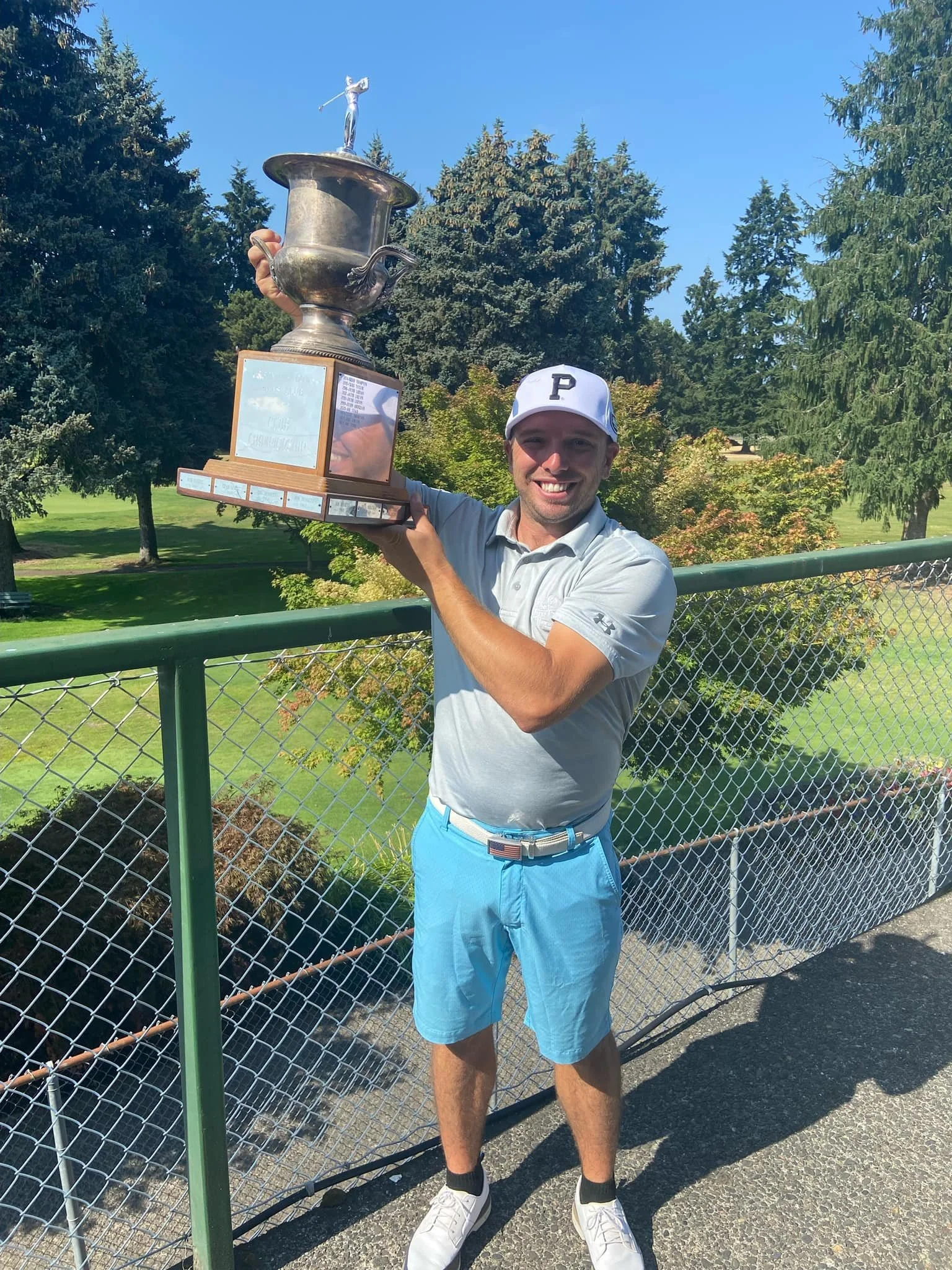 A man in a white golf shirt, light blue shorts, and a white cap with a black 'P' logo is smiling and holding up a large trophy with a figure of a golfer on top, outdoors near a green athletic field with trees in the background.