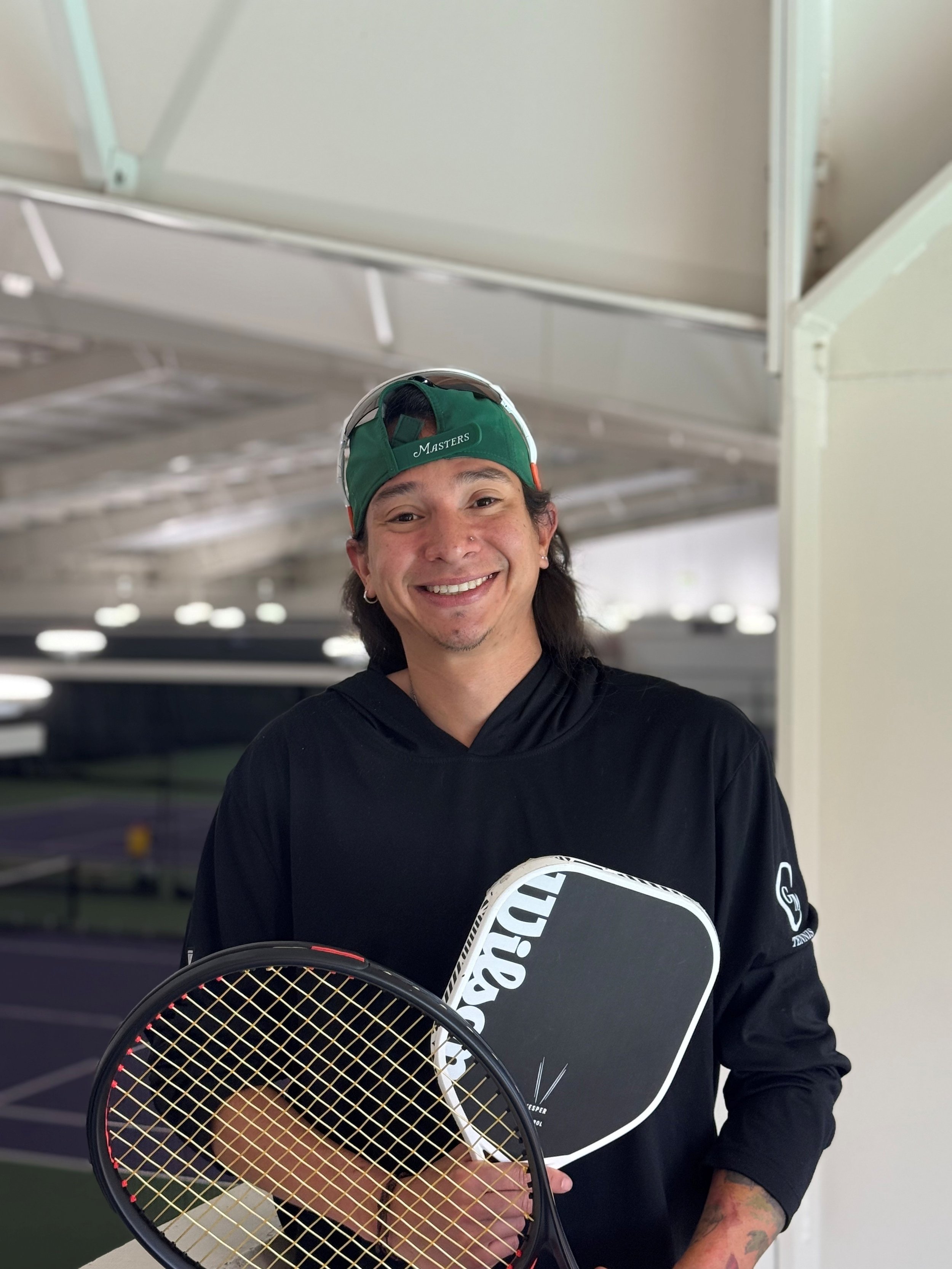 Smiling man holding a tennis racket and a paddle, wearing a black hoodie, a green cap, and sunglasses on his head, standing near an indoor tennis court.