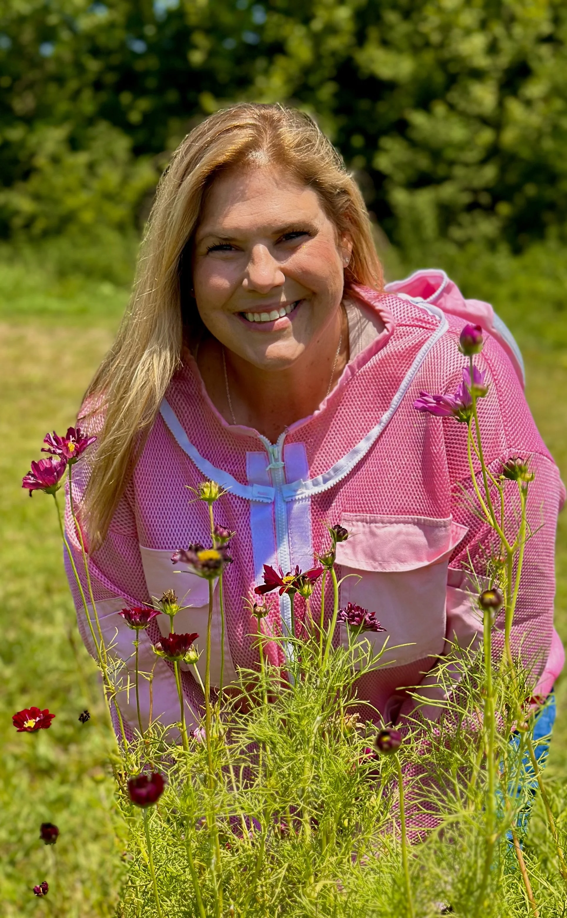A woman with long blonde hair smiling outdoors in a pink jacket, surrounded by blooming purple and pink flowers, with green trees in the background.