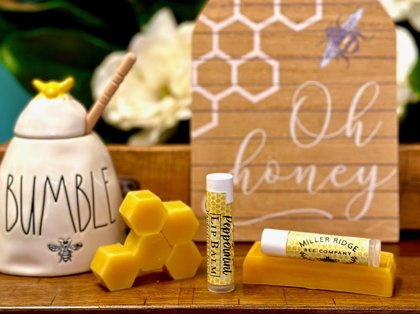 A display of honey-related products including a ceramic container labeled 'Bumble,' a white lip balm labeled 'Lip Balm,' a yellow honeycomb-shaped candle, and honey lip balm tubes from Miller Ridge Bee Company. There is a wooden sign in the background with the words 'Oh Honey' written in white cursive, and some white flowers and greenery behind.