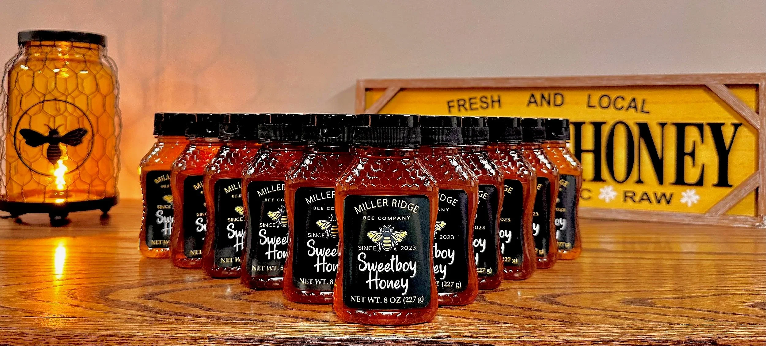 Multiple jars of Miller Ridge Bee Company Sweetboy Honey arranged on a wooden surface with a honey jar-shaped lamp featuring a bee design and a yellow sign reading 'Fresh and Local Honey Raw' in the background.