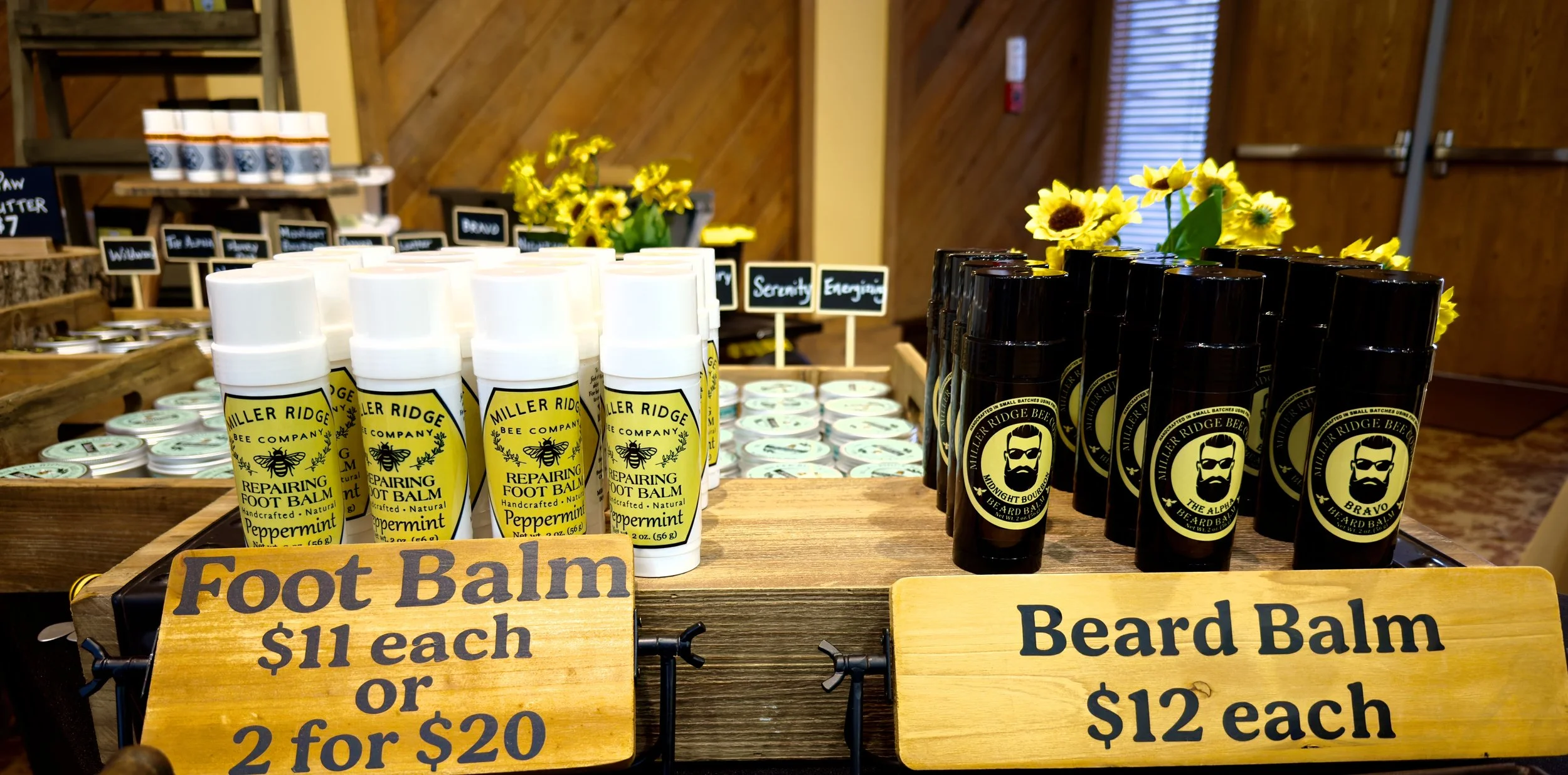 Display of Miller Ridge Bee Company foot balm and beard balm bottles on a wooden table with sunflower decorations and small chalkboard signs showing prices, in a shop or market setting.