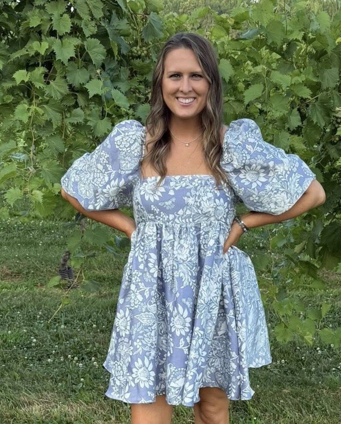 Woman standing outdoors in front of green leafy plants, wearing a light blue and white floral dress with puffy sleeves, smiling with her hands on her hips.