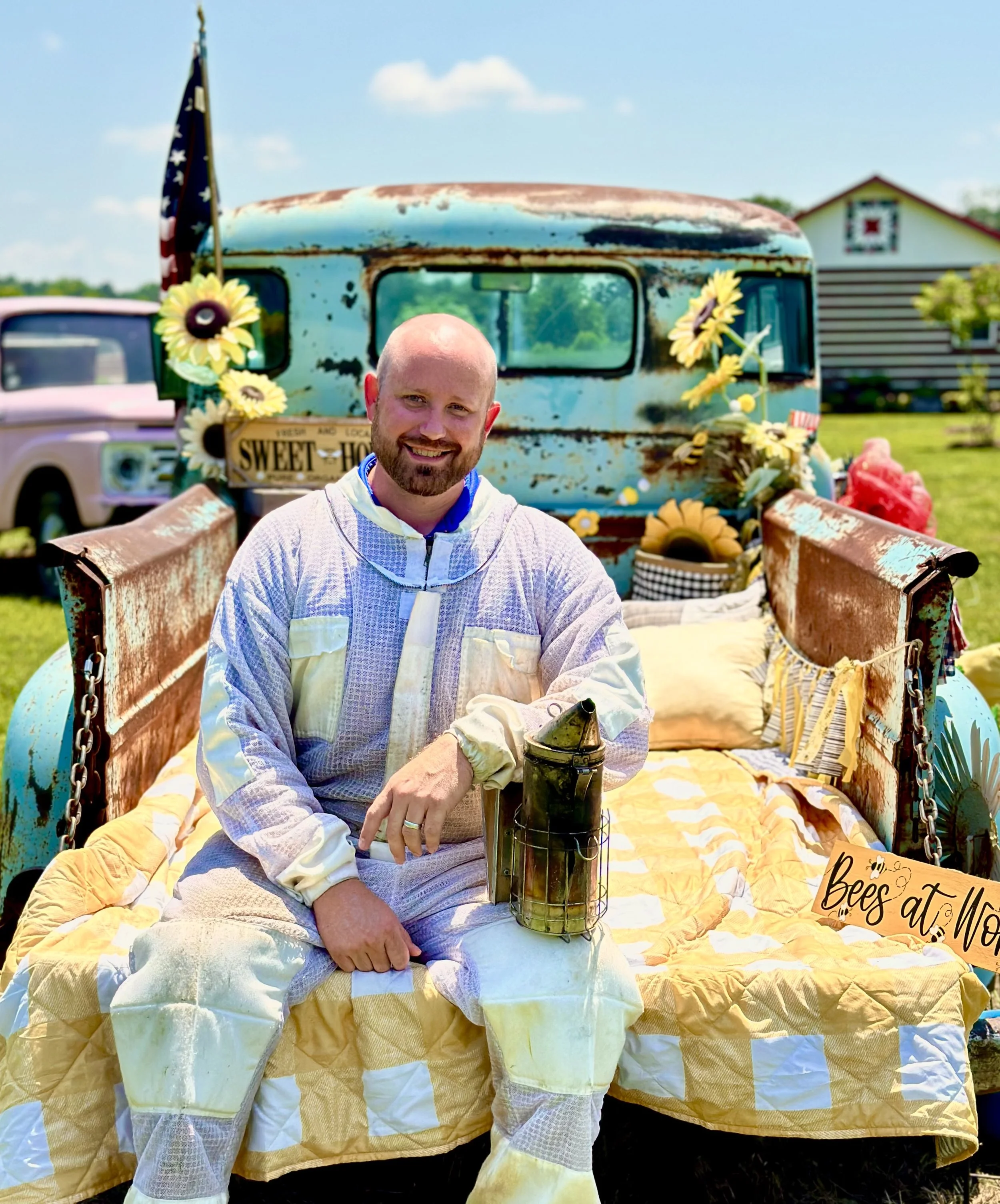 Erik smiling sitting on a bed of yellow and white quilt in a vintage truck bed decorated with sunflowers and a bees at work sign, outdoors with a house and other trucks in the background.