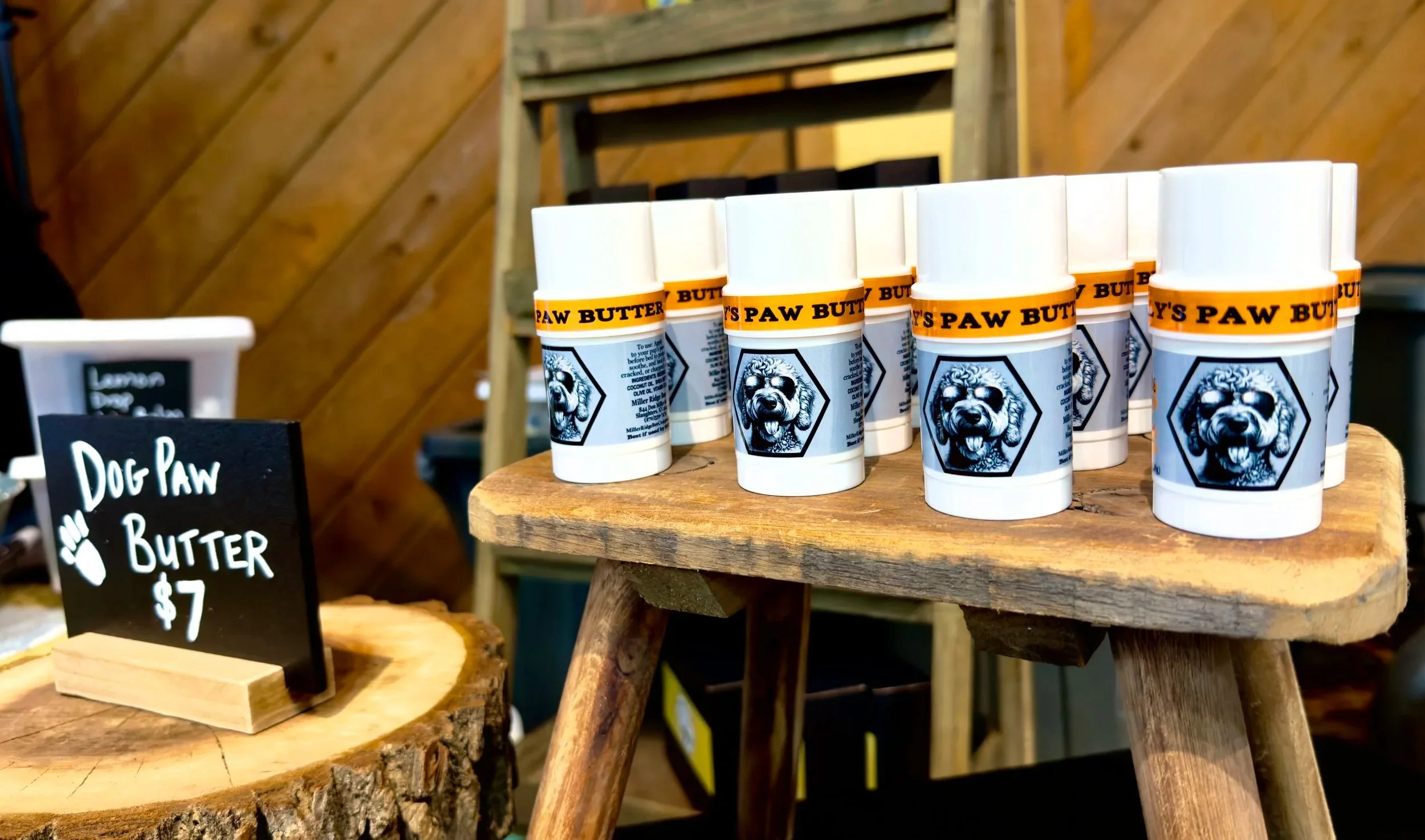 Containers of dog paw butter on a wooden table, with a small chalkboard sign in front that says 'Dog Paw Butter $7'.