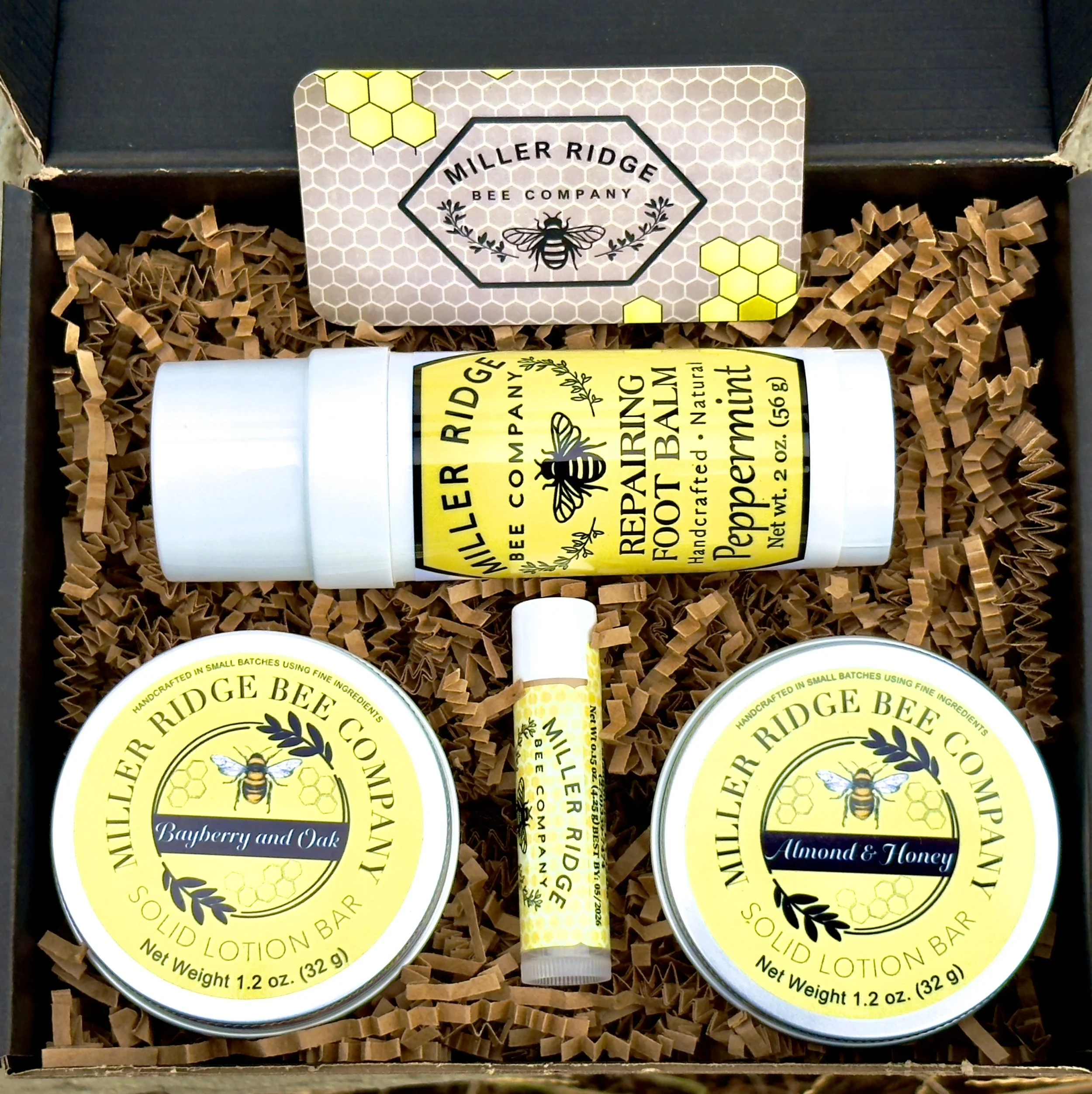 Gift box containing Miller Ridge Bee Company skincare products, including solid lotion bars in blueberry and oak, almond and honey flavors, a peppermint foot balm, and a bee-shaped lip balm, all arranged on brown shredded paper.