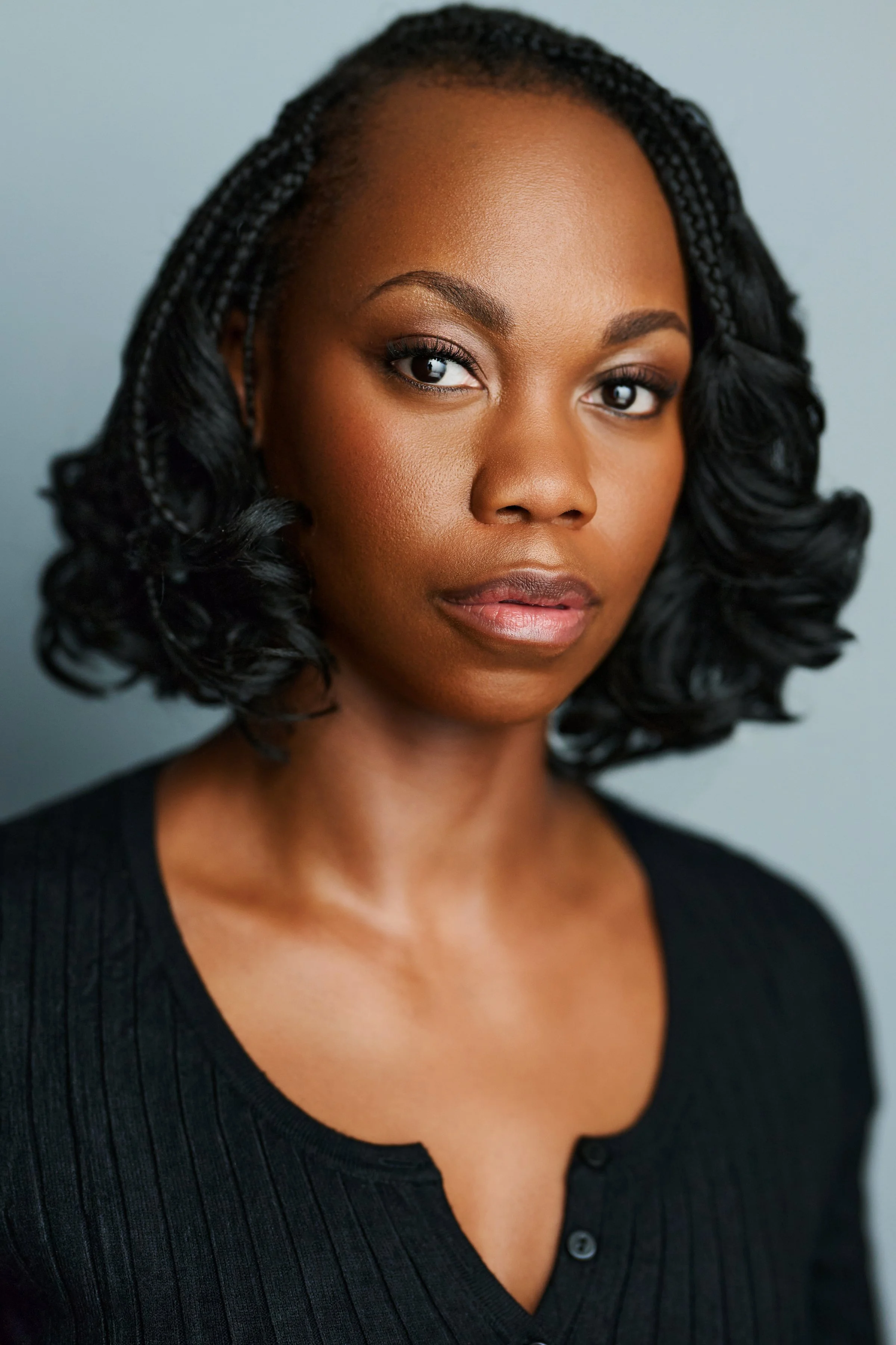 Portrait of a Black woman with shoulder-length curly hair, wearing a black top, looking directly at the camera with a serious expression.