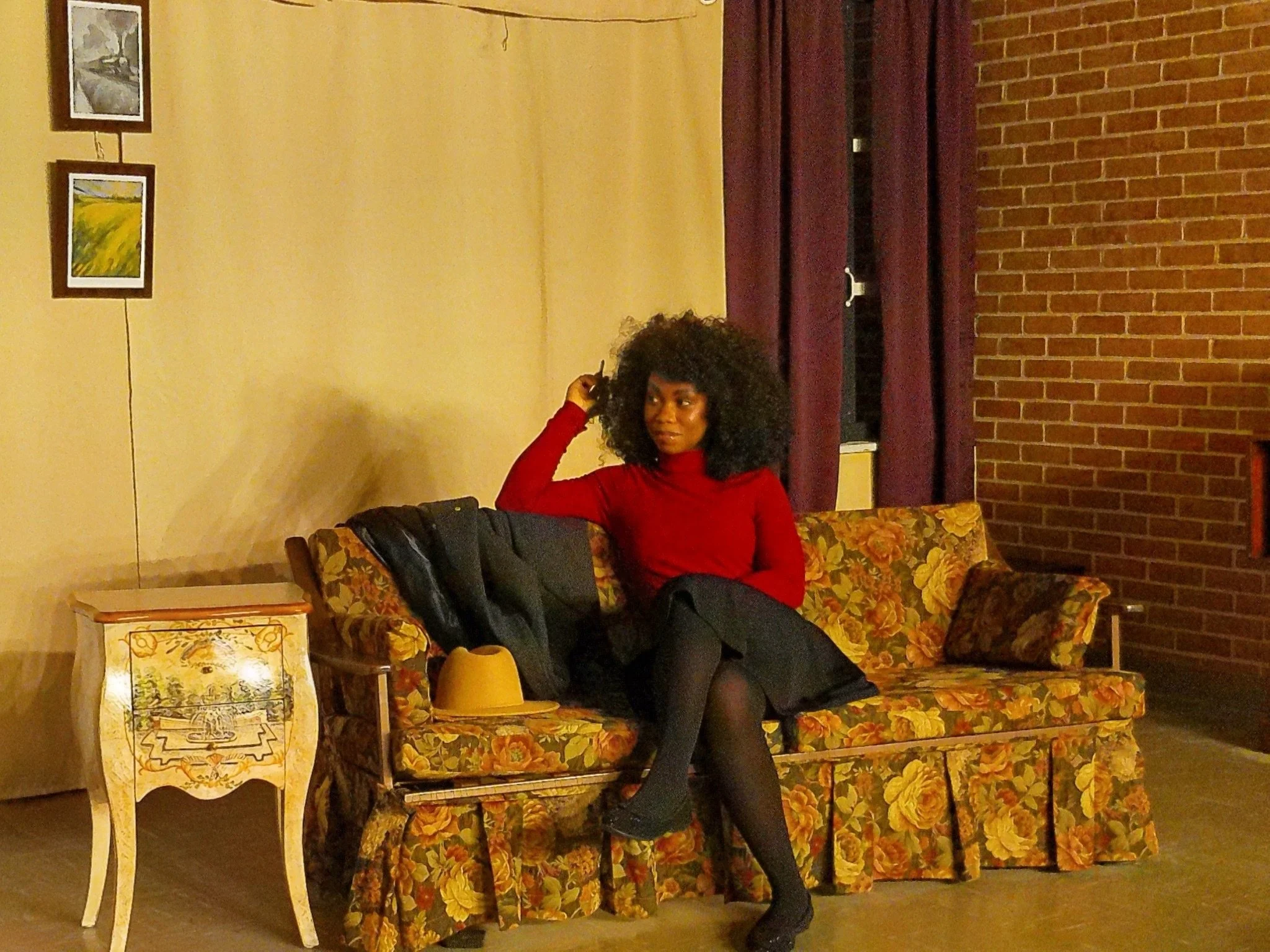 A woman with curly black hair wearing a red turtleneck, black skirt, and black tights sitting on a vintage floral sofa in a warmly lit room with brick wall and purple curtains.