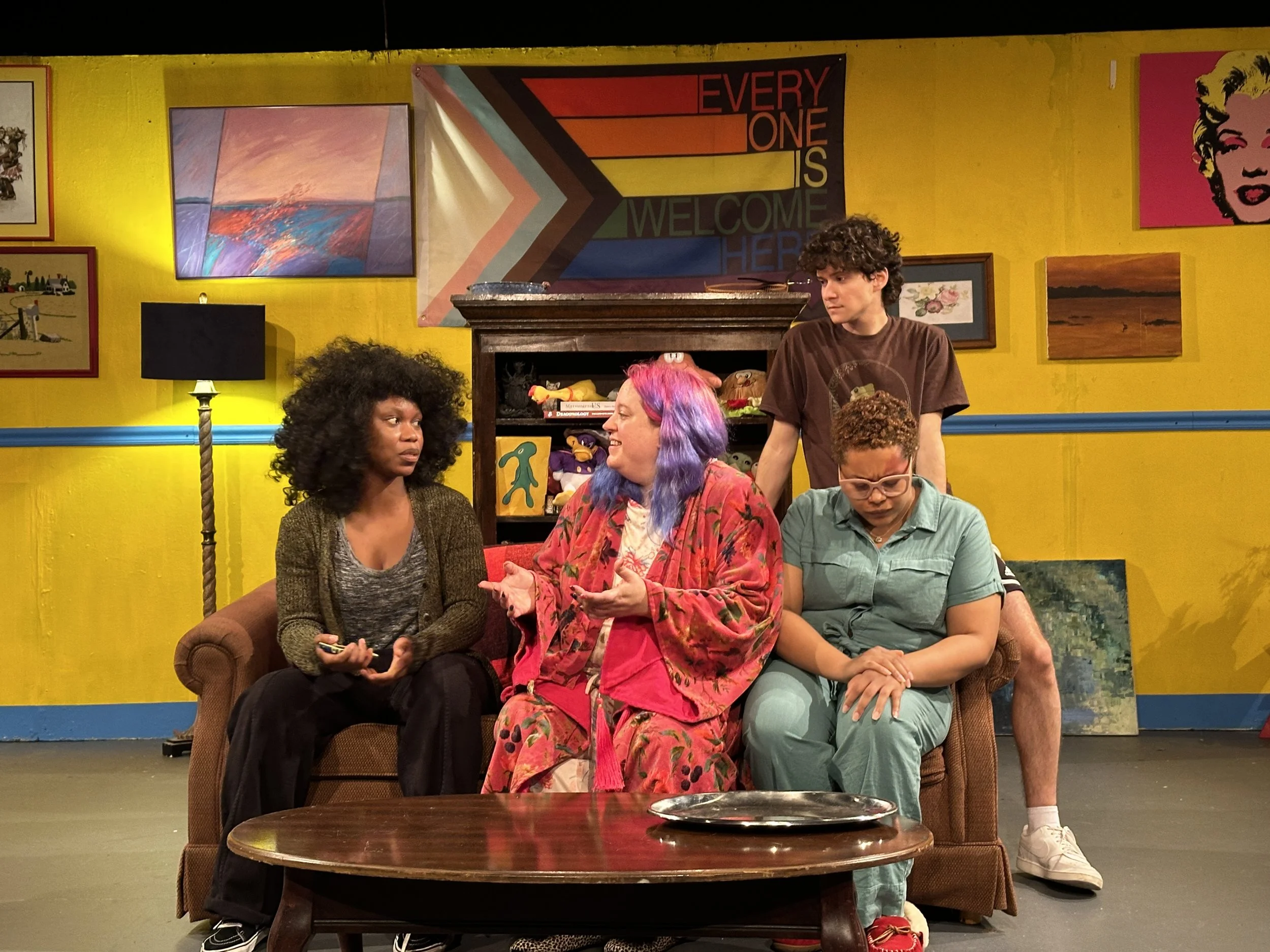 Four people sitting on a brown couch in a colorful room with yellow walls and artwork. One person with pink and purple hair speaking, others listening or looking down. Two standing behind the couch.