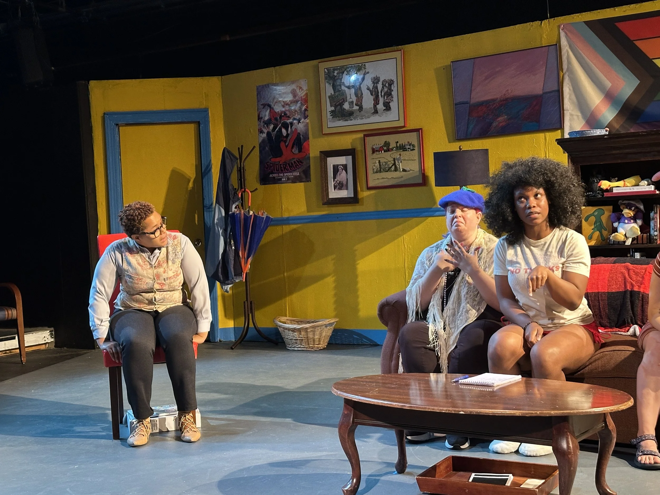 Three people are on stage in a theater set. The person on the left is sitting on a red chair, wearing glasses, a colorful vest, and dark pants. The two people on the right sit on a sofa; the person in the middle has short hair, a blue beret, and whit