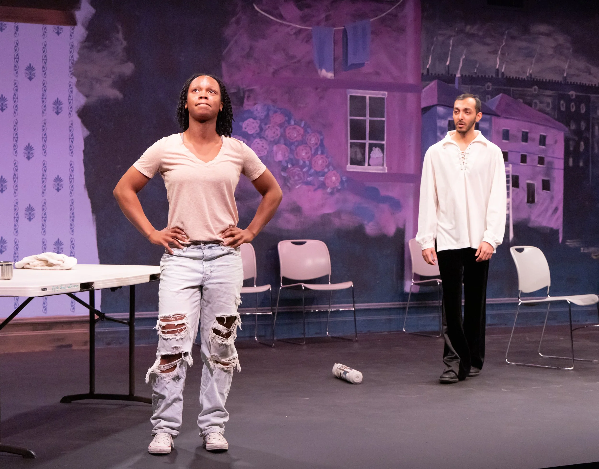 Two actors stand on stage during a play, with a painted backdrop of a house and flowers. The woman in the foreground has black hair, wearing a beige t-shirt and ripped jeans, with hands on her hips looking upward. The man in the background has short 