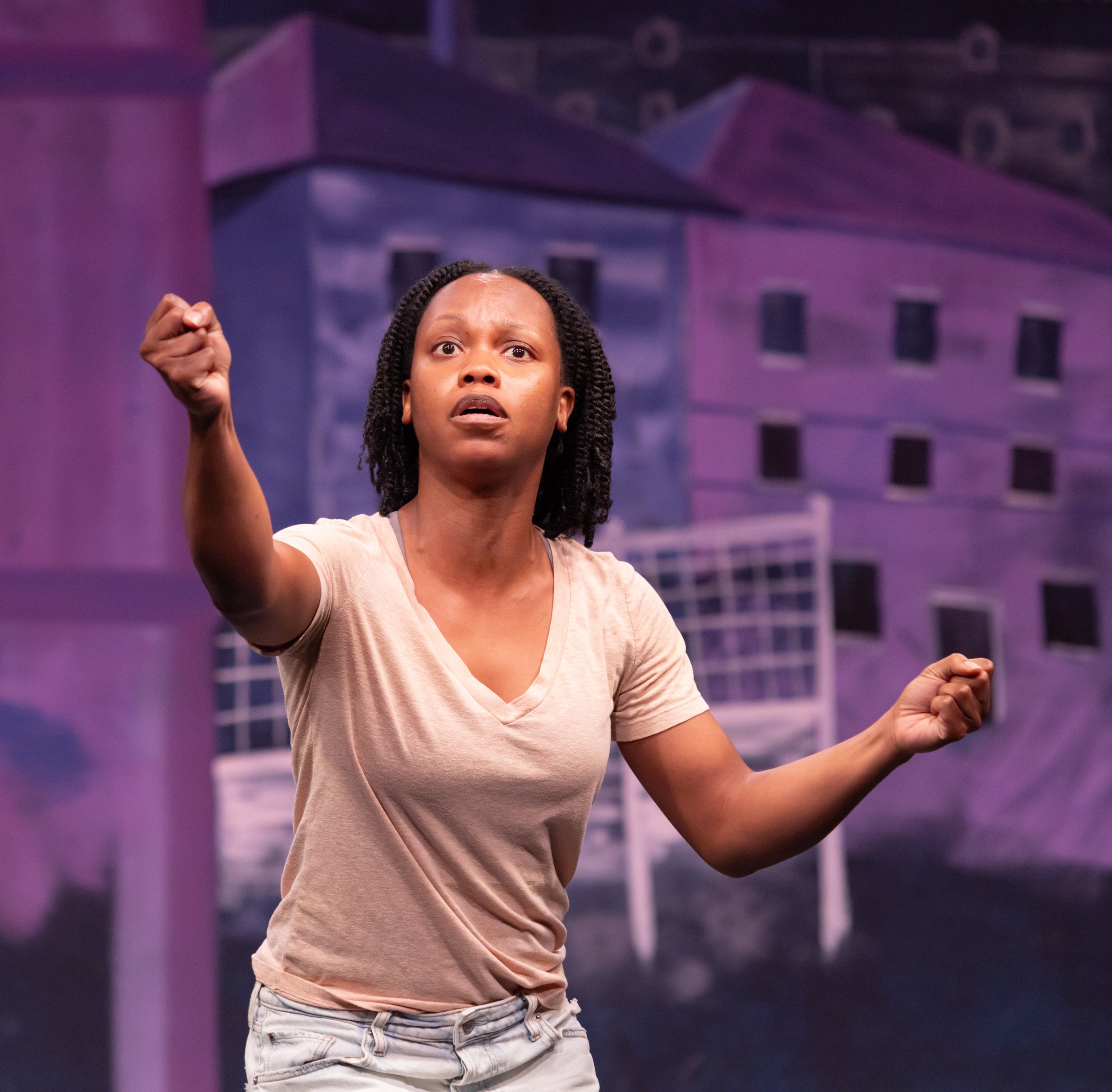 A woman with twisted hair and wearing a beige T-shirt appears to be acting with a distressed expression on her face, set against a painted backdrop of a purple building.