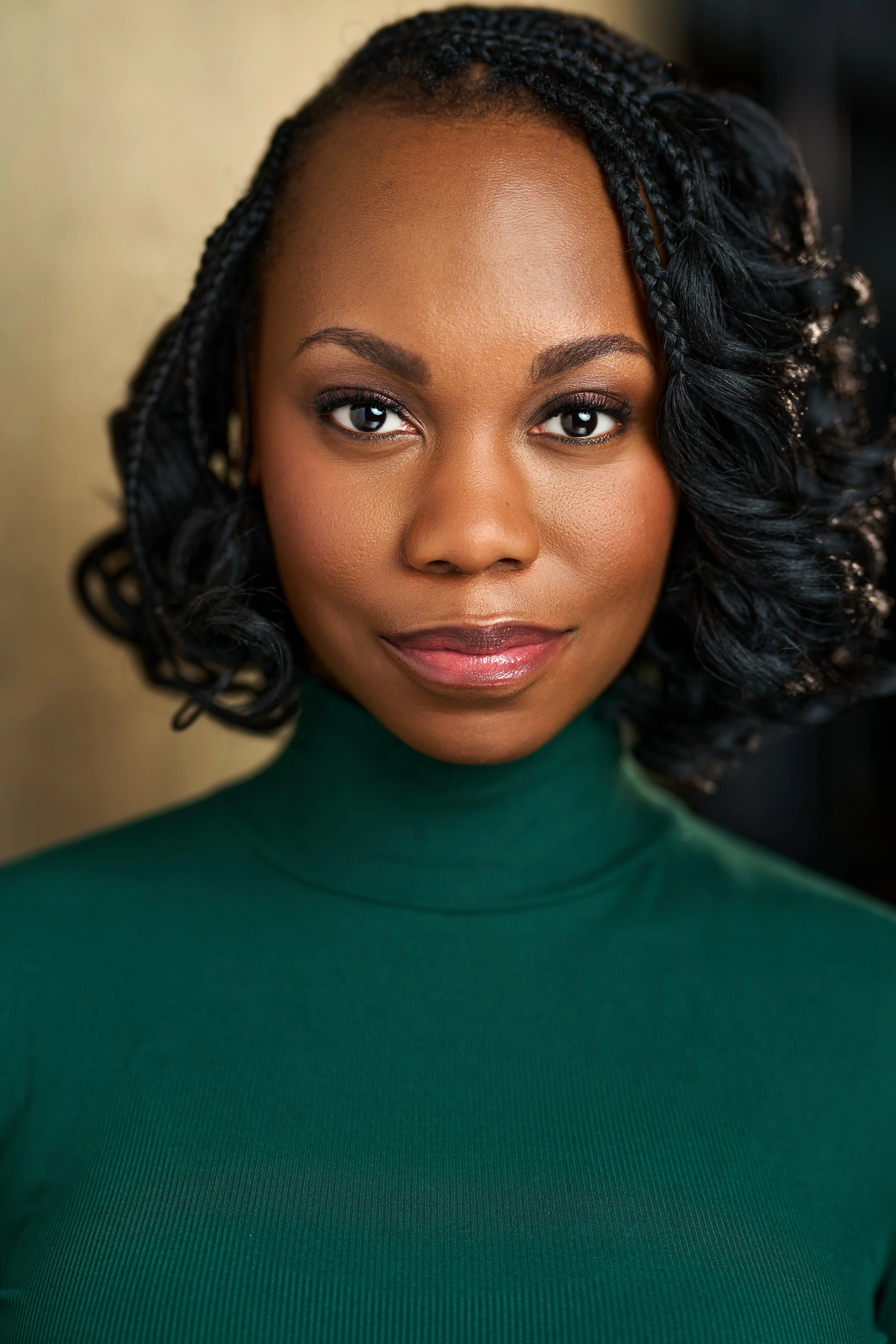 Close-up portrait of a young African-American woman with curly braided hair, wearing a green turtleneck, looking directly at the camera with a slight smile.